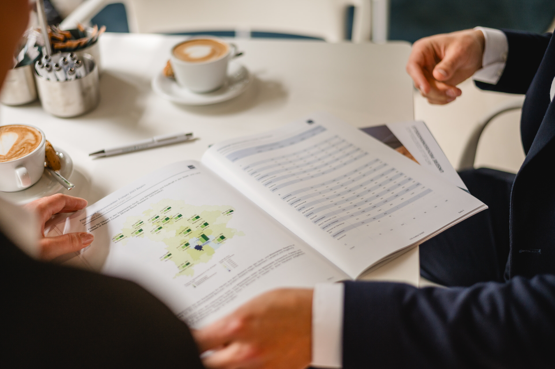 Christian Lorenz Fotograf Zwei Personen in Geschäftskleidung sitzen mit Kaffeetassen an einem Tisch und betrachten einen offenen Bericht mit Diagrammen und Karten. Nur ihre Hände und Arme sind sichtbar, während sie das Dokument besprechen.