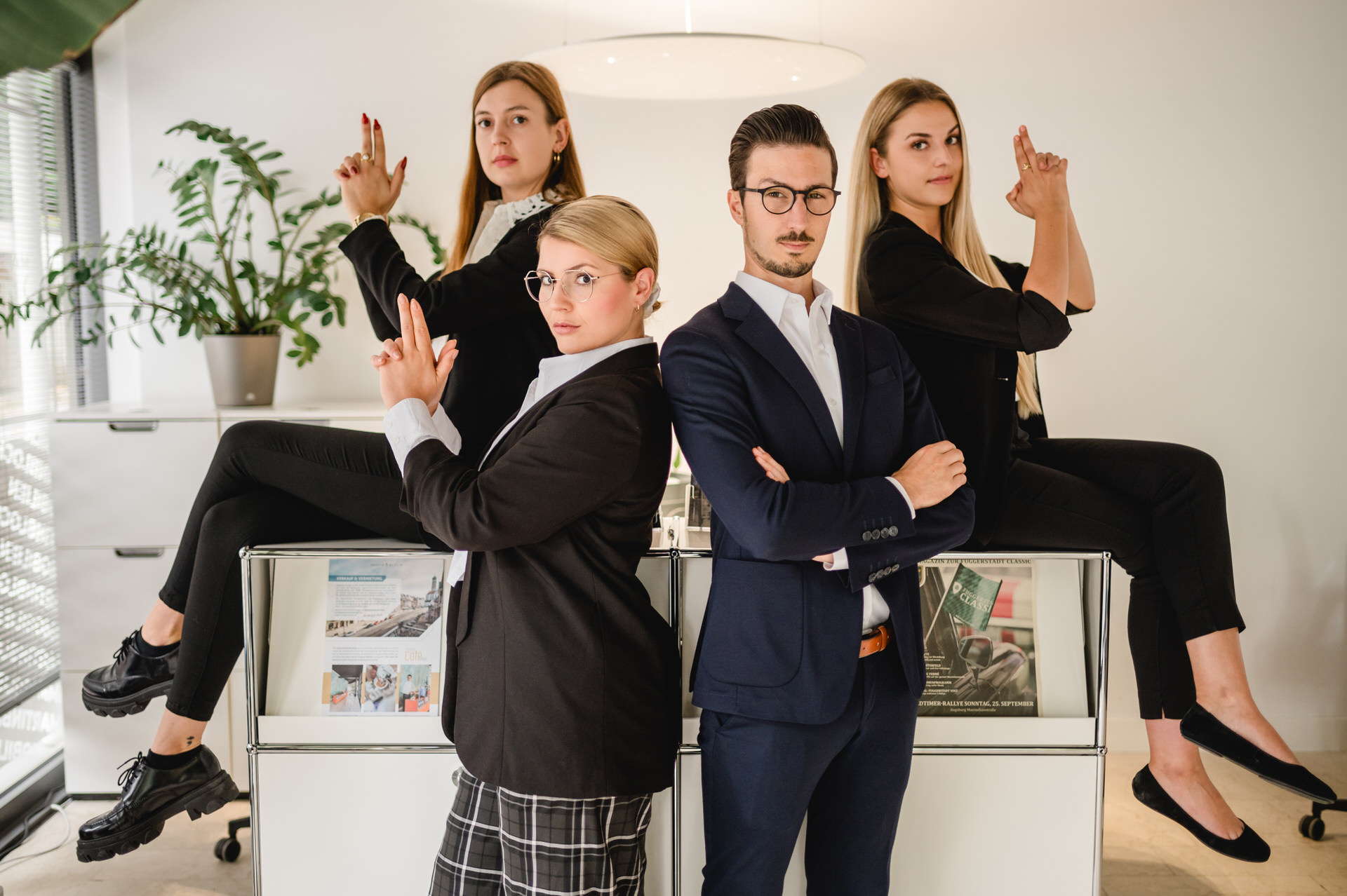 Christian Lorenz Fotograf Vier junge Berufstätige in Business-Kleidung posieren in einem Büro. Zwei Frauen sitzen mit Fingerpistolen auf den Schreibtischen, eine weitere Frau steht mit Fingerpistolen, und ein Mann steht mit verschränkten Armen davor, alle wirken selbstbewusst.