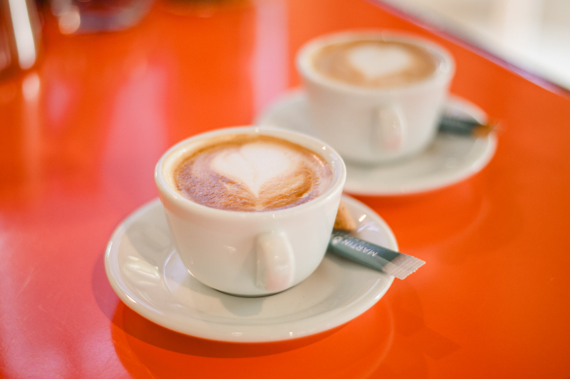 Christian Lorenz Fotograf Zwei weiße Cappuccinotassen mit herzförmigem Schaum sitzen auf weißen Untertassen auf einem leuchtend orangefarbenen Tisch. Jede Untertasse enthält ein kleines Zuckerpäckchen. Der Hintergrund ist sanft verschwommen.