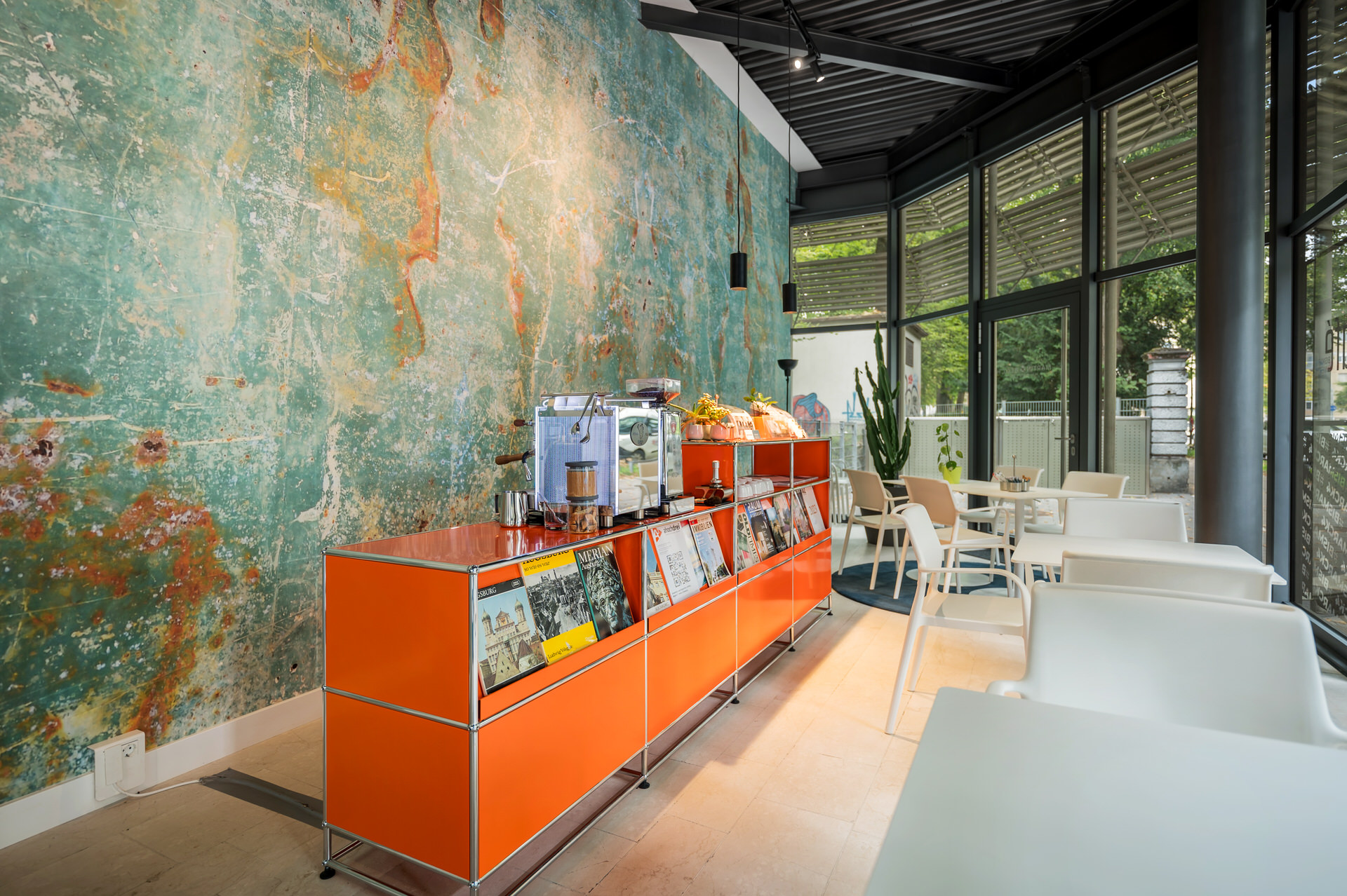 Christian Lorenz Fotograf Ein modernes Café mit einer Wand in Teal und Orange, einer orangefarbenen Theke mit Zeitschriften, Kaffeemaschinen und Snacks, weißen Tischen und Stühlen, großen Fenstern und natürlichem Licht.