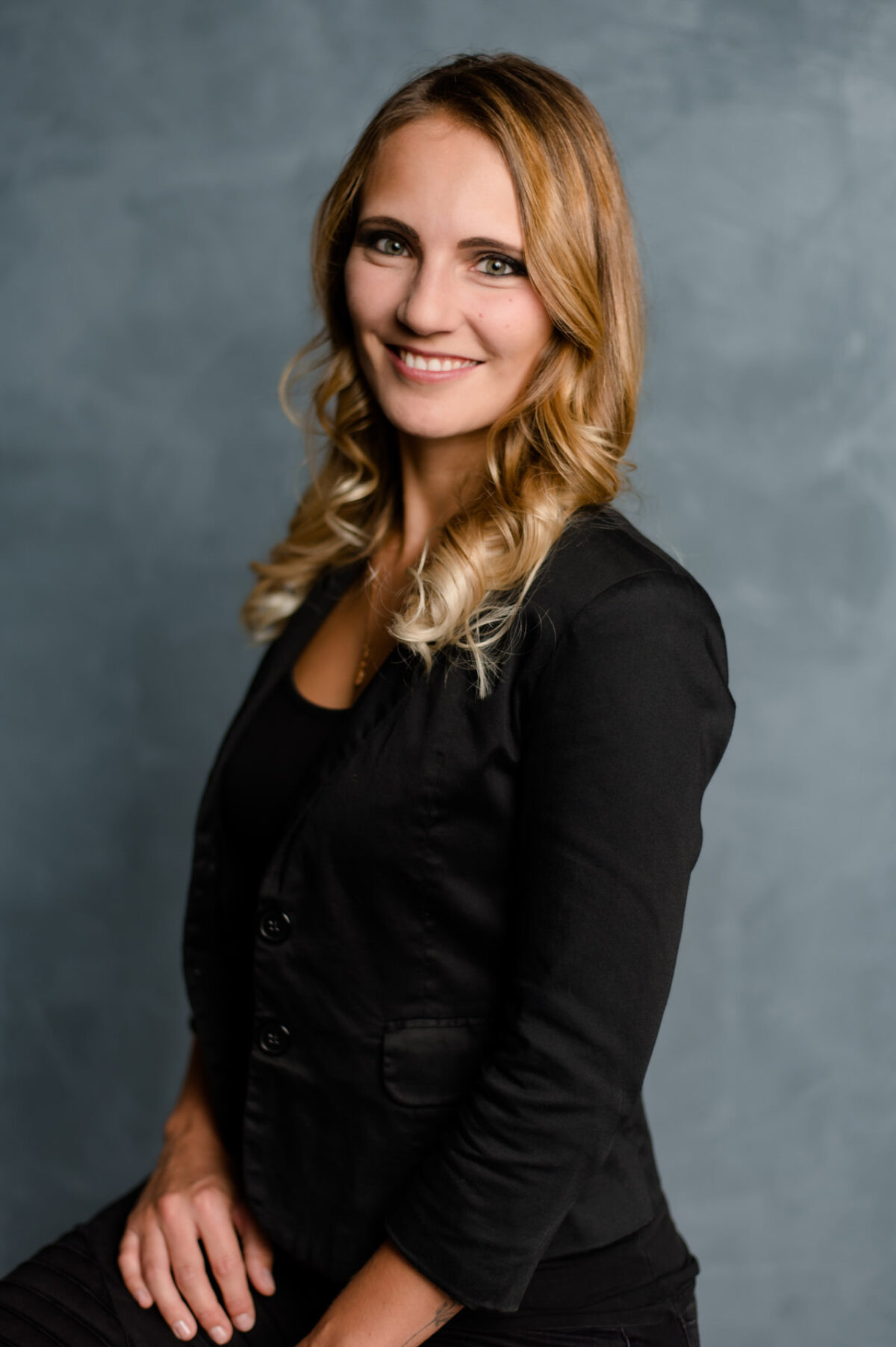 Christian Lorenz Fotograf Eine Frau mit langem, gewelltem blondem Haar, die einen schwarzen Blazer über einem schwarzen Oberteil trägt, lächelt in die Kamera vor einem grauen Studiohintergrund. Christian Lorenz Fotograf Eine Frau mit langem, gewelltem blondem Haar, die einen schwarzen Blazer über einem schwarzen Oberteil trägt, lächelt in die Kamera vor einem grauen Studiohintergrund.