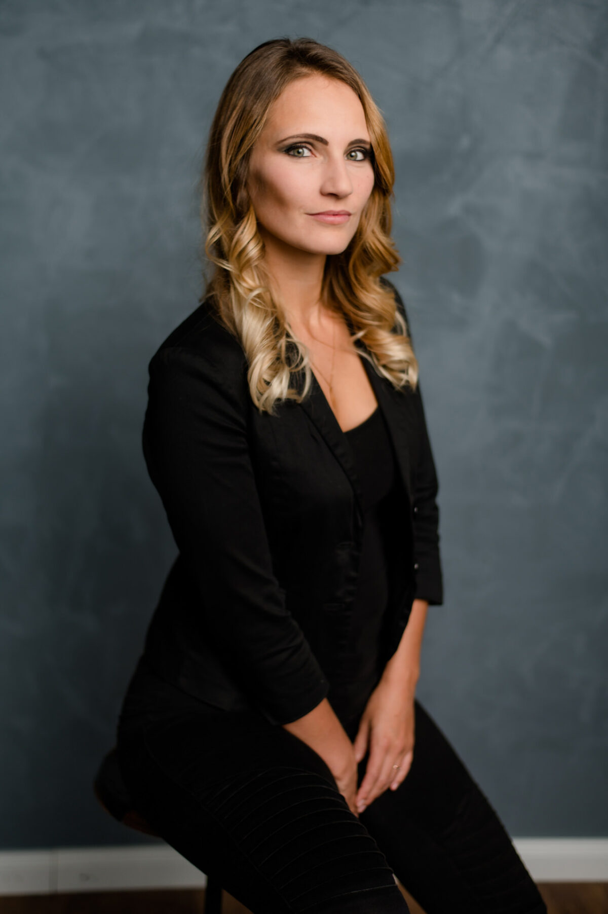 Christian Lorenz Fotograf Eine Frau mit langem, gewelltem blondem Haar sitzt auf einem Hocker vor einem strukturierten grauen Hintergrund, trägt einen schwarzen Blazer und eine Hose und schaut selbstbewusst in die Kamera. Christian Lorenz Fotograf Eine Frau mit langem, gewelltem blondem Haar sitzt auf einem Hocker vor einem strukturierten grauen Hintergrund, trägt einen schwarzen Blazer und eine Hose und schaut selbstbewusst in die Kamera.