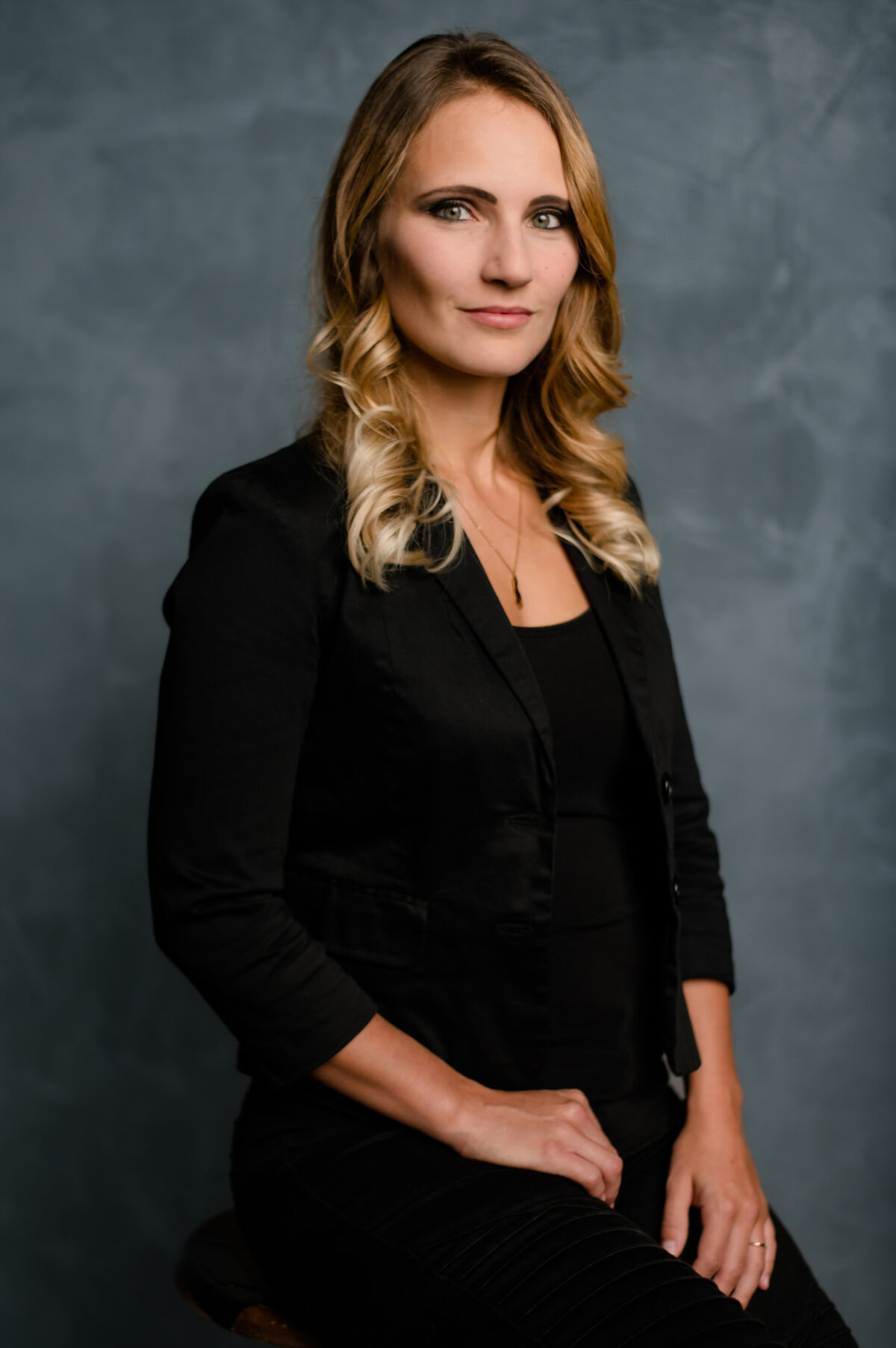 Christian Lorenz Fotograf Eine Frau mit langem, gewelltem blondem Haar sitzt auf einem Hocker vor einem grauen Hintergrund. Sie trägt einen schwarzen Blazer über einem schwarzen Oberteil und blickt ruhig und mit neutralem Blick in die Kamera. Christian Lorenz Fotograf Eine Frau mit langem, gewelltem blondem Haar sitzt auf einem Hocker vor einem grauen Hintergrund. Sie trägt einen schwarzen Blazer über einem schwarzen Oberteil und blickt ruhig und mit neutralem Blick in die Kamera.