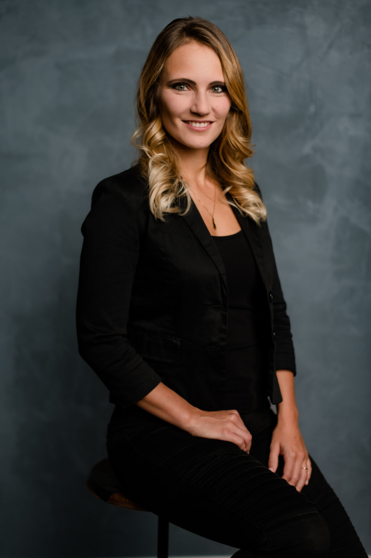 Christian Lorenz Fotograf Eine Frau mit langem, gewelltem blondem Haar, die einen schwarzen Blazer und eine Hose trägt, sitzt auf einem Hocker vor einem strukturierten dunkelgrauen Hintergrund und lächelt in die Kamera. Christian Lorenz Fotograf Eine Frau mit langem, gewelltem blondem Haar, die einen schwarzen Blazer und eine Hose trägt, sitzt auf einem Hocker vor einem strukturierten dunkelgrauen Hintergrund und lächelt in die Kamera.