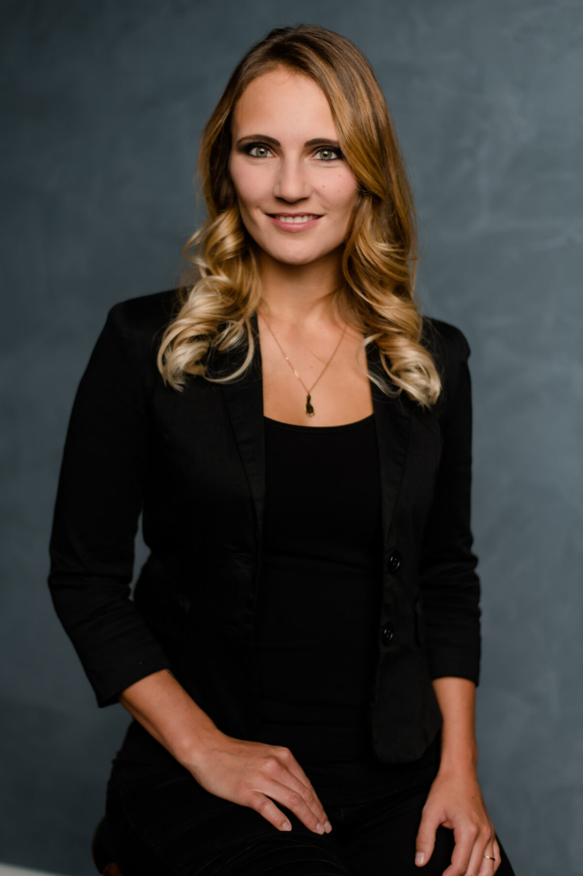 Christian Lorenz Fotograf Eine Frau mit blondem, gewelltem Haar, die einen schwarzen Blazer und ein schwarzes Oberteil trägt, sitzt vor einem grauen, strukturierten Hintergrund, lächelt leicht und hat die Hände in den Schoß gelegt. Christian Lorenz Fotograf Eine Frau mit blondem, gewelltem Haar, die einen schwarzen Blazer und ein schwarzes Oberteil trägt, sitzt vor einem grauen, strukturierten Hintergrund, lächelt leicht und hat die Hände in den Schoß gelegt.