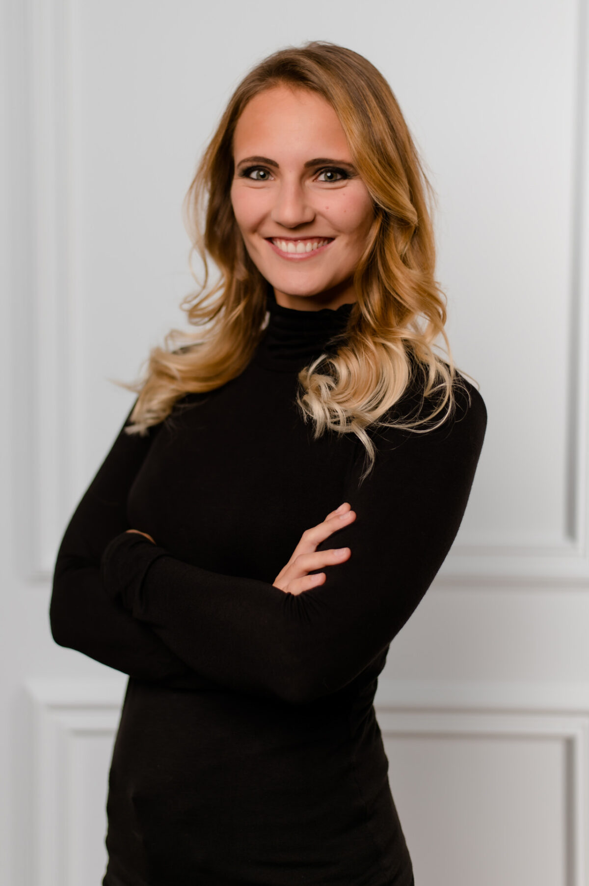 Christian Lorenz Fotograf Eine Frau mit langen, gewellten blonden Haaren und einem schwarzen Rollkragenpullover steht mit verschränkten Armen und einem selbstbewussten Lächeln vor einem weißen, getäfelten Hintergrund. Christian Lorenz Fotograf Eine Frau mit langen, gewellten blonden Haaren und einem schwarzen Rollkragenpullover steht mit verschränkten Armen und einem selbstbewussten Lächeln vor einem weißen, getäfelten Hintergrund.