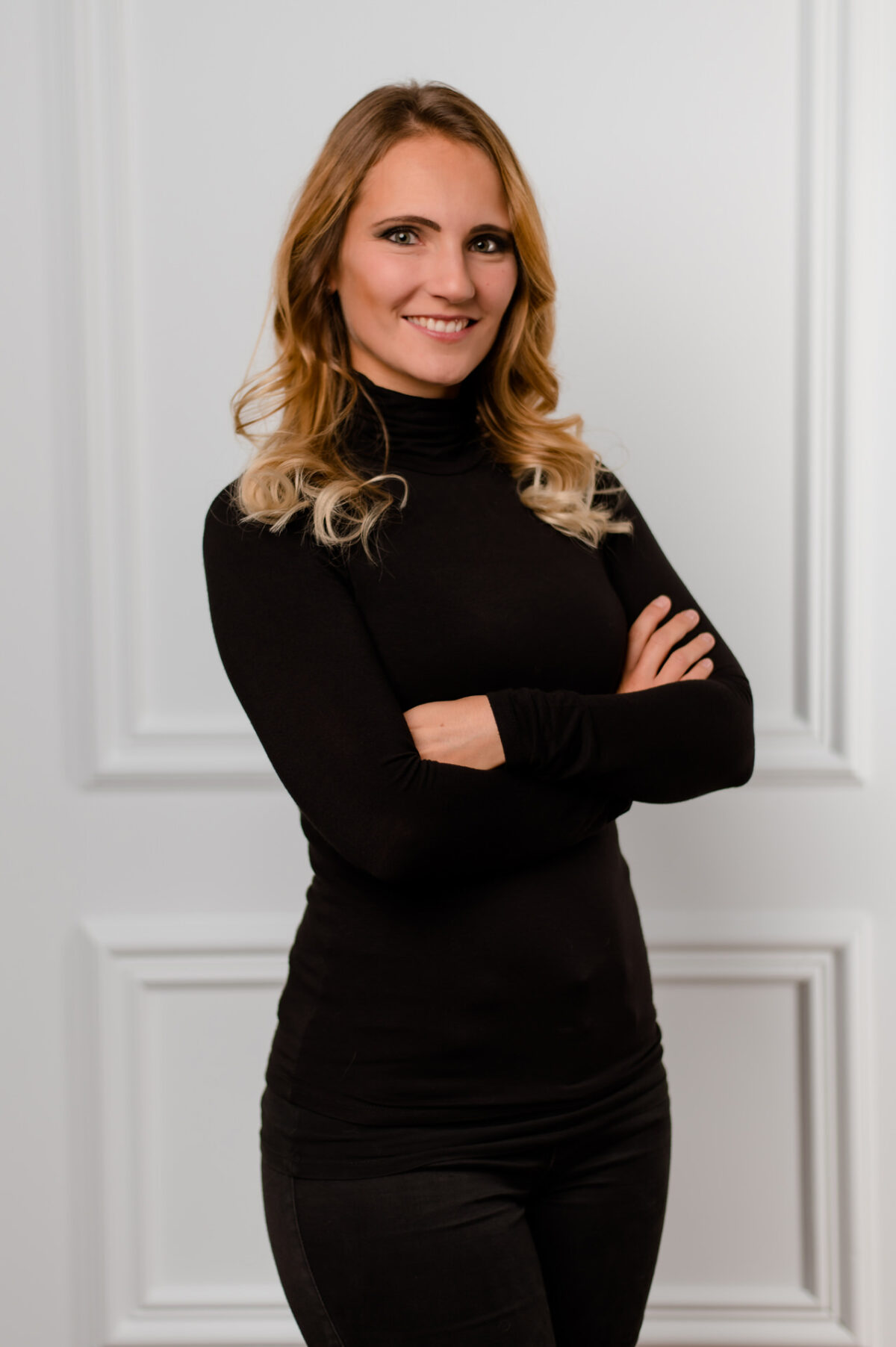 Christian Lorenz Fotograf Eine Frau mit langen blonden Haaren und einem schwarzen Rollkragenpullover steht mit verschränkten Armen lächelnd vor einer weiß getäfelten Wand. Christian Lorenz Fotograf Eine Frau mit langen blonden Haaren und einem schwarzen Rollkragenpullover steht mit verschränkten Armen lächelnd vor einer weiß getäfelten Wand.