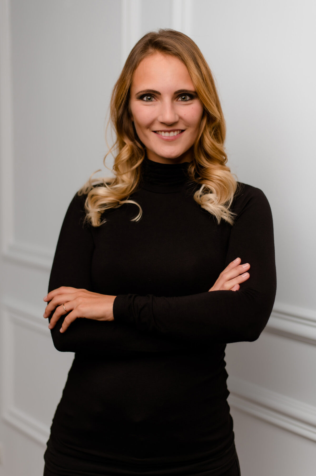 Christian Lorenz Fotograf Eine Frau mit langen, gewellten blonden Haaren und einem schwarzen Rollkragenpullover steht mit verschränkten Armen und einem selbstbewussten Lächeln vor einer weiß getäfelten Wand. Christian Lorenz Fotograf Eine Frau mit langen, gewellten blonden Haaren und einem schwarzen Rollkragenpullover steht mit verschränkten Armen und einem selbstbewussten Lächeln vor einer weiß getäfelten Wand.