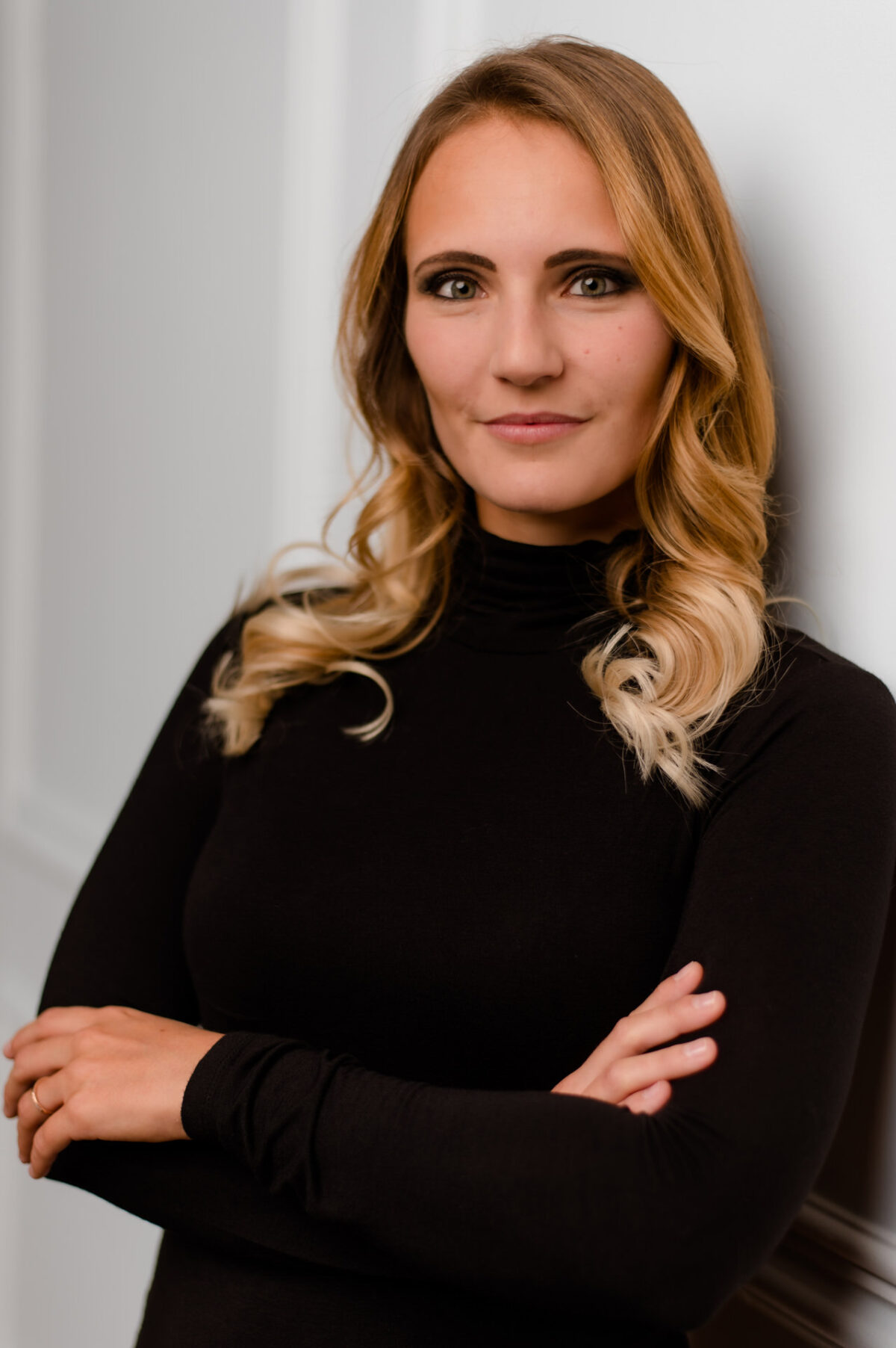 Christian Lorenz Fotograf Eine selbstbewusste Frau mit langen blonden Haaren in lockeren Locken und einem schwarzen Rollkragenpullover steht mit verschränkten Armen und lächelt leicht. Sie ist gegen eine helle Wand gelehnt. Christian Lorenz Fotograf Eine selbstbewusste Frau mit langen blonden Haaren in lockeren Locken und einem schwarzen Rollkragenpullover steht mit verschränkten Armen und lächelt leicht. Sie ist gegen eine helle Wand gelehnt.