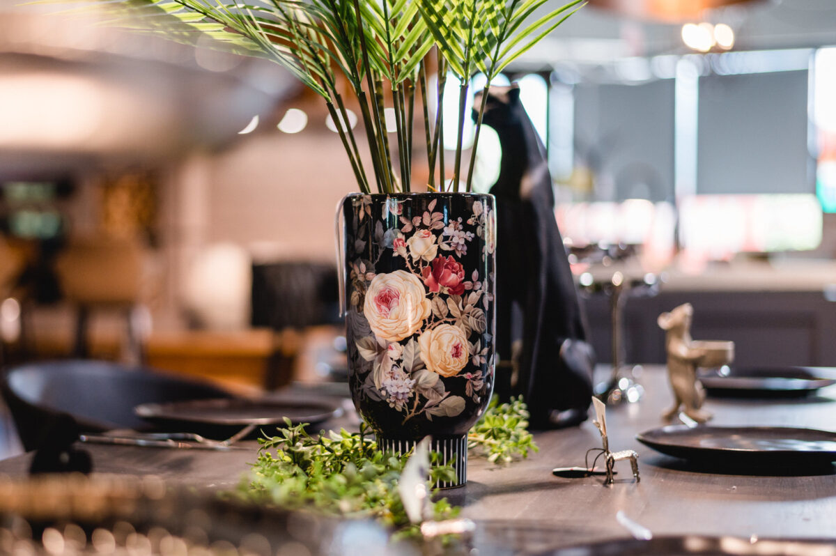 Christian Lorenz Fotograf Nahaufnahme einer Blumenvase mit grünen Palmenblättern, die auf einem dekorierten Esstisch mit schwarzen Tellern und tierförmigen Dekorationsgegenständen in einem modernen, sanft beleuchteten Raum steht. Christian Lorenz Fotograf Nahaufnahme einer Blumenvase mit grünen Palmenblättern, die auf einem dekorierten Esstisch mit schwarzen Tellern und tierförmigen Dekorationsgegenständen in einem modernen, sanft beleuchteten Raum steht.