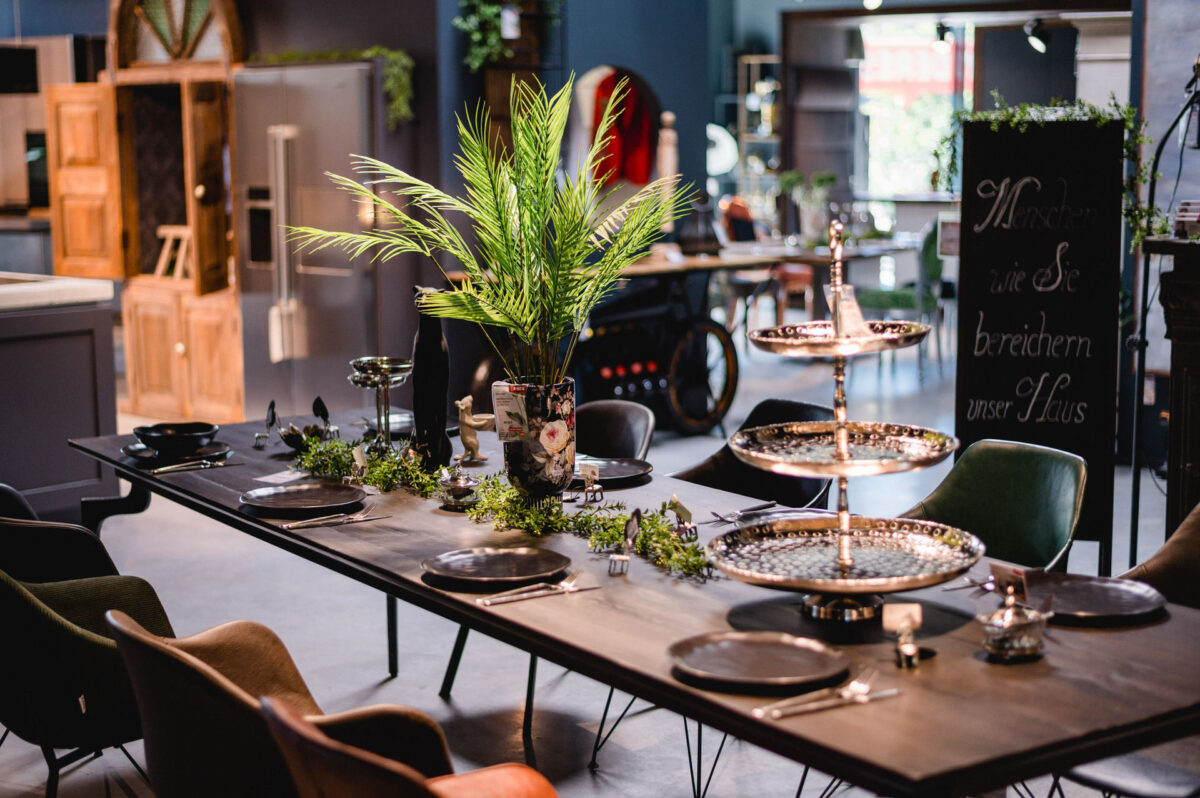 Christian Lorenz Fotograf Ein modern gedeckter Esstisch mit schwarzen Tellern, silbernem Besteck, Grünzeug, einer hohen Vase mit Palmenblättern und verzierten, gestaffelten Tabletts steht in einem stilvollen, offenen Raum mit eklektischem Dekor. Christian Lorenz Fotograf Ein modern gedeckter Esstisch mit schwarzen Tellern, silbernem Besteck, Grünzeug, einer hohen Vase mit Palmenblättern und verzierten, gestaffelten Tabletts steht in einem stilvollen, offenen Raum mit eklektischem Dekor.