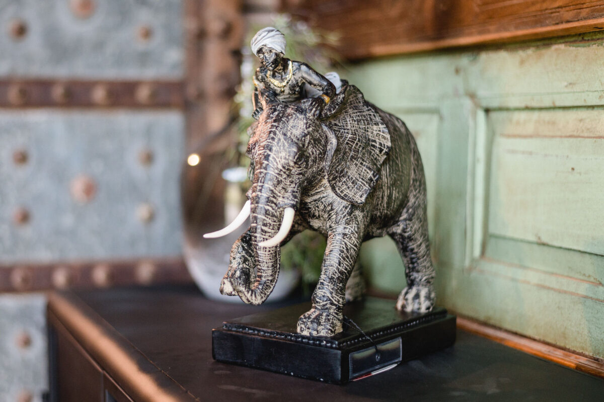 Christian Lorenz Fotograf Eine dekorative Skulptur eines Elefanten mit großen Stoßzähnen und einem Reiter, der einen Turban trägt, steht auf einem schwarzen Sockel und wird in einem Innenraum vor einer grün-braunen Wand aufgestellt. Christian Lorenz Fotograf Eine dekorative Skulptur eines Elefanten mit großen Stoßzähnen und einem Reiter, der einen Turban trägt, steht auf einem schwarzen Sockel und wird in einem Innenraum vor einer grün-braunen Wand aufgestellt.