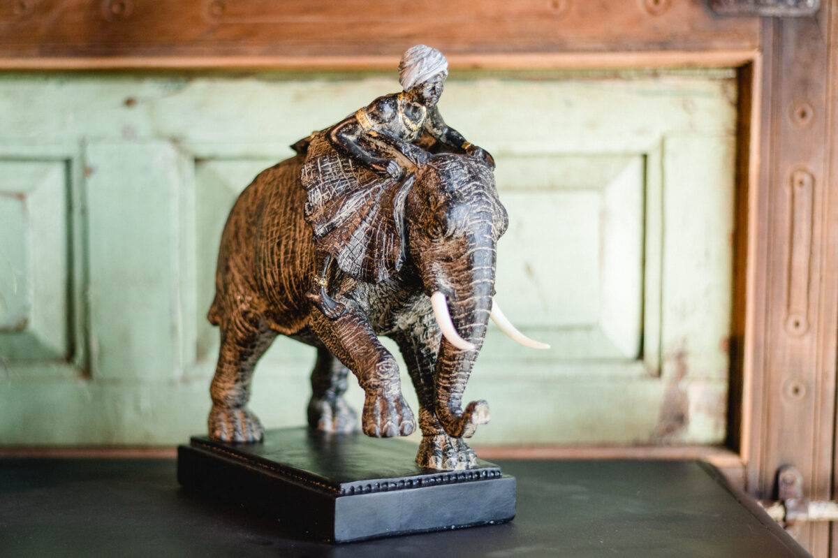 Christian Lorenz Fotograf Eine detaillierte Skulptur eines Elefanten mit einem Reiter auf dem Rücken, die auf einem schwarzen, rechteckigen Sockel vor einem rustikalen, grün-braunen Holzhintergrund steht. Christian Lorenz Fotograf Eine detaillierte Skulptur eines Elefanten mit einem Reiter auf dem Rücken, die auf einem schwarzen, rechteckigen Sockel vor einem rustikalen, grün-braunen Holzhintergrund steht.