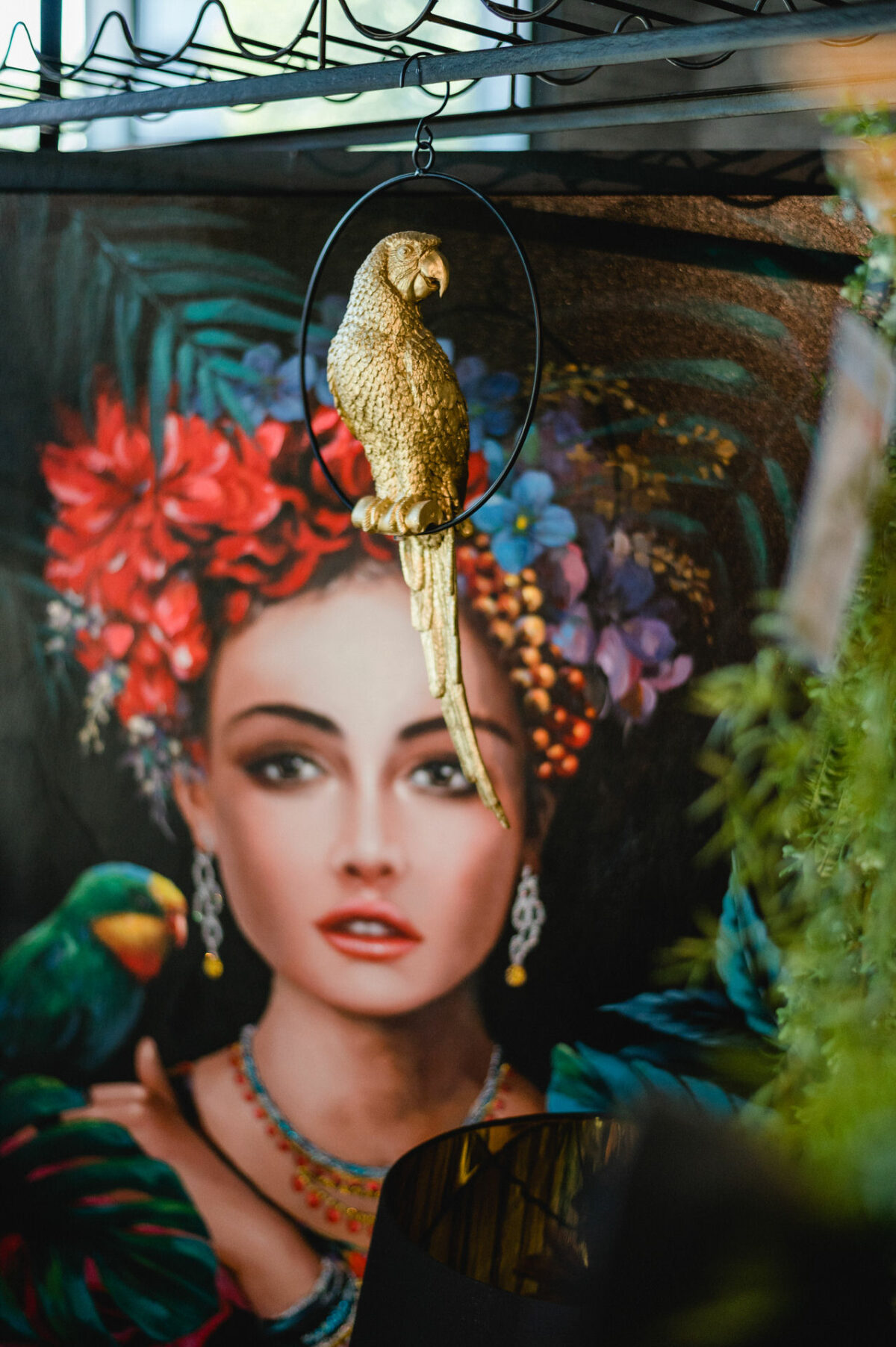 Christian Lorenz Fotograf Eine dekorative goldene Papageienfigur hängt vor einem lebendigen Gemälde einer Frau mit Blumen im Haar und einem grünen Papagei auf der Schulter. Der Hintergrund ist bunt und üppig mit Pflanzen. Christian Lorenz Fotograf Eine dekorative goldene Papageienfigur hängt vor einem lebendigen Gemälde einer Frau mit Blumen im Haar und einem grünen Papagei auf der Schulter. Der Hintergrund ist bunt und üppig mit Pflanzen.