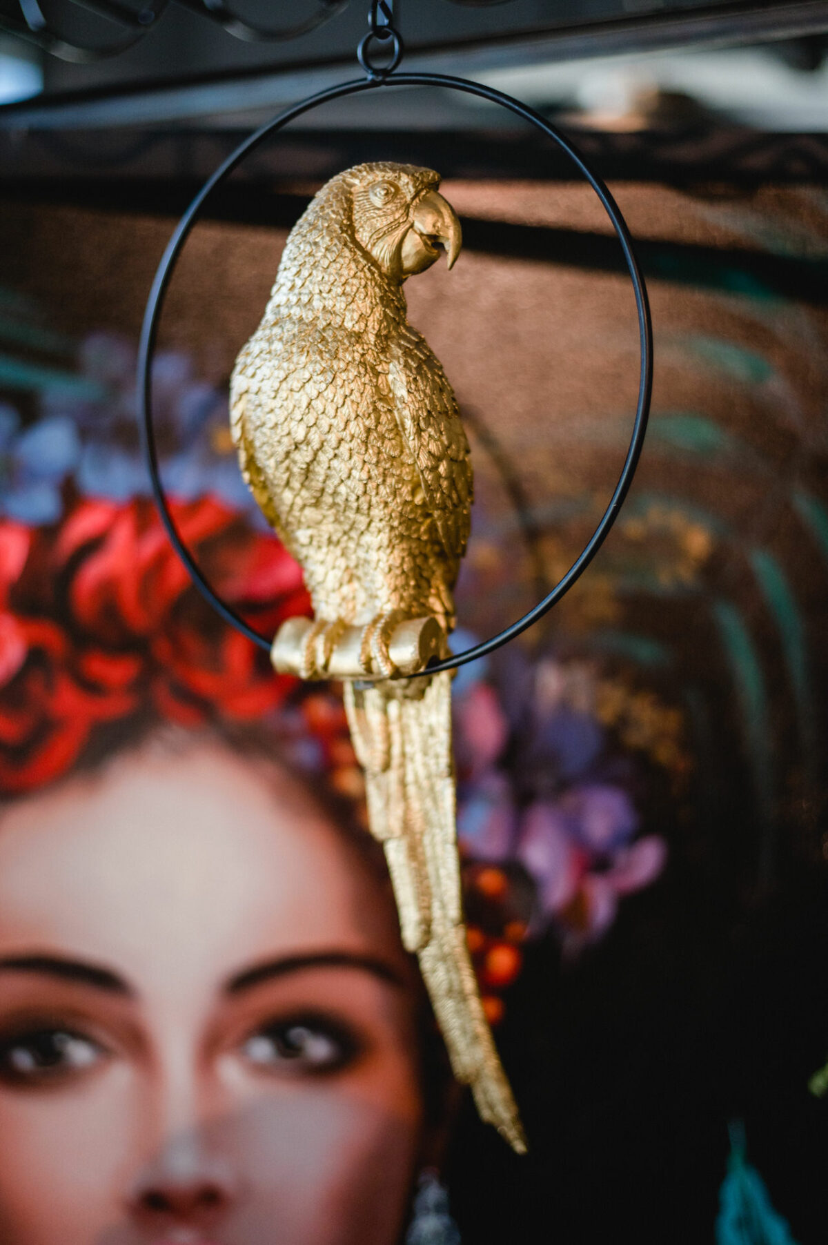 Christian Lorenz Fotograf Eine goldene Papageienstatue, die auf einem runden schwarzen Ring sitzt, mit einem verschwommenen farbigen Porträt eines Frauengesichts und einer geblümten Kopfbedeckung im Hintergrund. Christian Lorenz Fotograf Eine goldene Papageienstatue, die auf einem runden schwarzen Ring sitzt, mit einem verschwommenen farbigen Porträt eines Frauengesichts und einer geblümten Kopfbedeckung im Hintergrund.