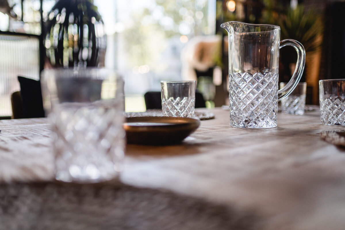 Christian Lorenz Fotograf Ein klarer Glaskrug und passende Gläser mit Rautenmuster stehen auf einem Holztisch in einem sanft beleuchteten Raum mit verschwommenen Hintergrundelementen und Sonnenlicht, das durch die Fenster fällt. Christian Lorenz Fotograf Ein klarer Glaskrug und passende Gläser mit Rautenmuster stehen auf einem Holztisch in einem sanft beleuchteten Raum mit verschwommenen Hintergrundelementen und Sonnenlicht, das durch die Fenster fällt.