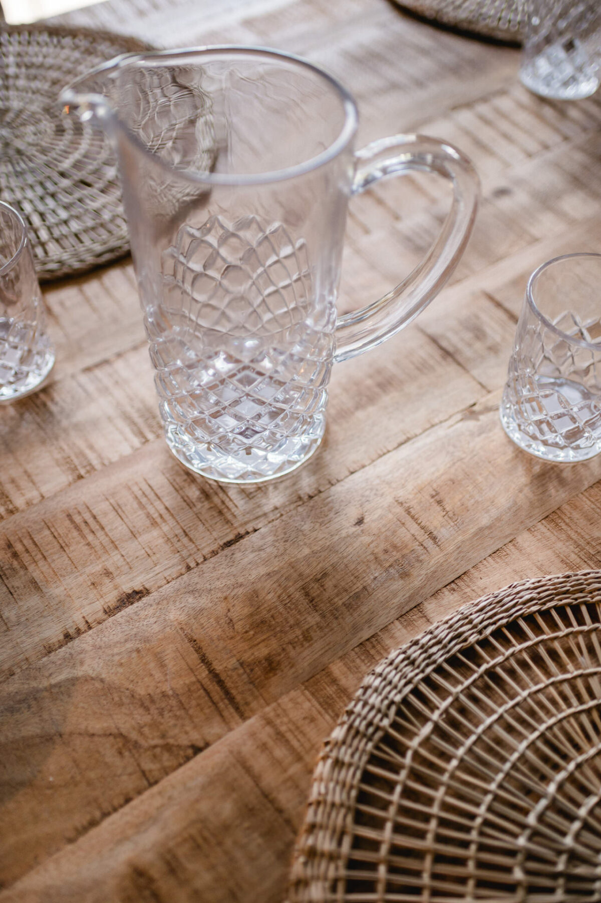 Christian Lorenz Fotograf Ein klarer Glaskrug und zwei passende Gläser mit einem strukturierten Rautenmuster stehen auf einem Holztisch mit runden, geflochtenen Platzsets. Die Tischoberfläche ist rustikal und hell gefärbt. Christian Lorenz Fotograf Ein klarer Glaskrug und zwei passende Gläser mit einem strukturierten Rautenmuster stehen auf einem Holztisch mit runden, geflochtenen Platzsets. Die Tischoberfläche ist rustikal und hell gefärbt.