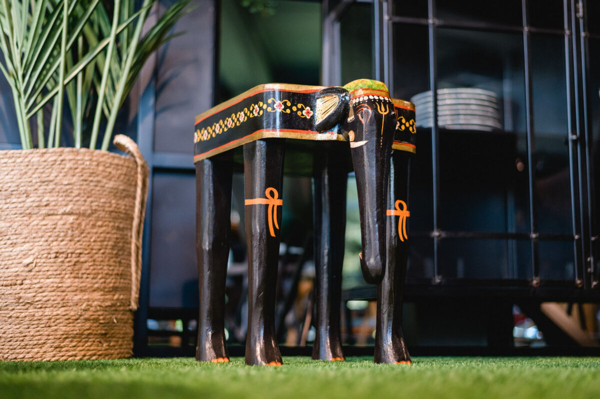 Christian Lorenz Fotograf Ein kleiner schwarzer Holztisch mit Beinen in Form eines Elefanten, verziert mit orangefarbenen und goldenen Mustern, steht auf einem grünen Teppich neben einem großen geflochtenen Korb mit hohen grünen Pflanzen. Christian Lorenz Fotograf Ein kleiner schwarzer Holztisch mit Beinen in Form eines Elefanten, verziert mit orangefarbenen und goldenen Mustern, steht auf einem grünen Teppich neben einem großen geflochtenen Korb mit hohen grünen Pflanzen.