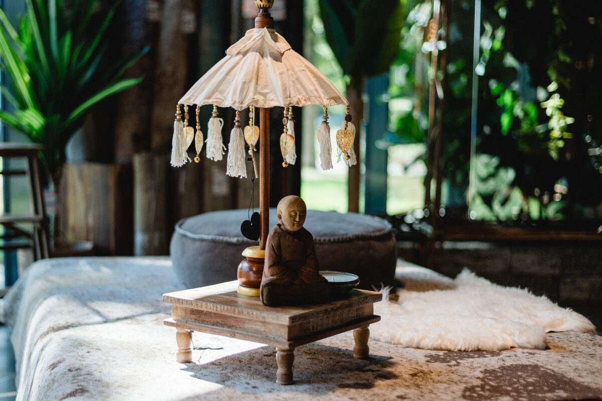 Christian Lorenz Fotograf Eine kleine Buddha-Statue sitzt auf einem Holzpodest unter einem dekorativen Schirm mit hängenden Ornamenten, der auf einem Tisch mit sanfter natürlicher Beleuchtung und Grün im Hintergrund steht. Christian Lorenz Fotograf Eine kleine Buddha-Statue sitzt auf einem Holzpodest unter einem dekorativen Schirm mit hängenden Ornamenten, der auf einem Tisch mit sanfter natürlicher Beleuchtung und Grün im Hintergrund steht.