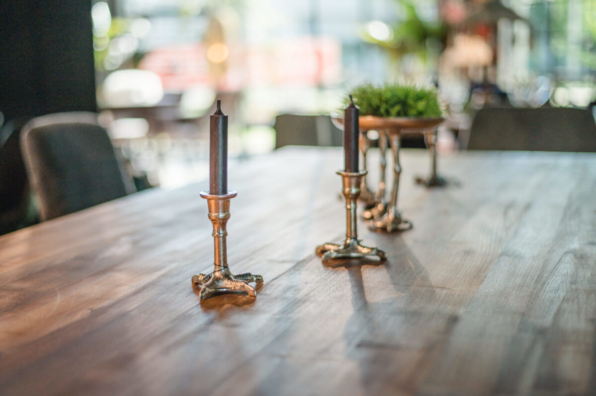 Christian Lorenz Fotograf Ein Holztisch mit drei verschnörkelten Kerzenhaltern aus Messing, zwei davon mit unbeleuchteten dunklen Kerzen, und eine kleine Topfpflanze im Hintergrund. Die Umgebung scheint ein Innenraum mit natürlichem Licht zu sein. Christian Lorenz Fotograf Ein Holztisch mit drei verschnörkelten Kerzenhaltern aus Messing, zwei davon mit unbeleuchteten dunklen Kerzen, und eine kleine Topfpflanze im Hintergrund. Die Umgebung scheint ein Innenraum mit natürlichem Licht zu sein.