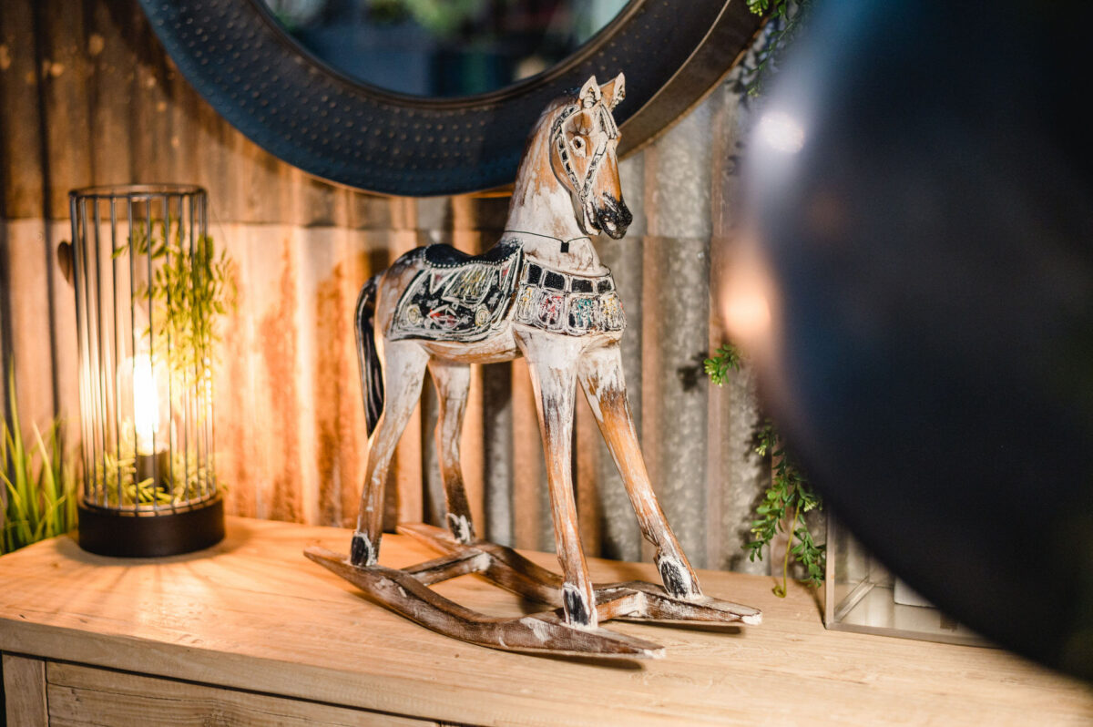 Christian Lorenz Fotograf Ein dekoratives hölzernes Schaukelpferd mit verschlungenen Mustern steht auf einem hellen Holztisch neben einer beleuchteten Laterne mit Grünzeug darin und schafft eine gemütliche, rustikale Atmosphäre. Christian Lorenz Fotograf Ein dekoratives hölzernes Schaukelpferd mit verschlungenen Mustern steht auf einem hellen Holztisch neben einer beleuchteten Laterne mit Grünzeug darin und schafft eine gemütliche, rustikale Atmosphäre.