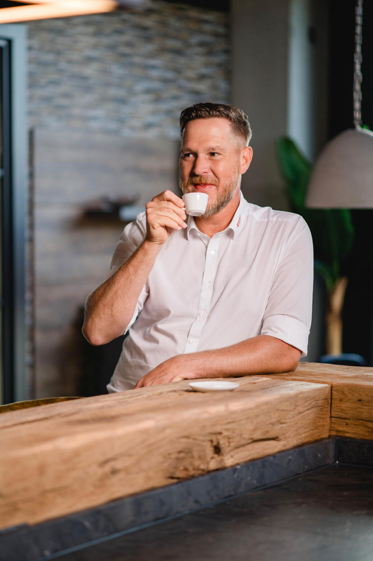 Christian Lorenz Fotograf Ein Mann in einem weißen Hemd steht an einem hölzernen Tresen, lächelt und hält eine kleine weiße Tasse Kaffee an seinen Mund in einem modernen, warm beleuchteten Innenraum. Christian Lorenz Fotograf Ein Mann in einem weißen Hemd steht an einem hölzernen Tresen, lächelt und hält eine kleine weiße Tasse Kaffee an seinen Mund in einem modernen, warm beleuchteten Innenraum.