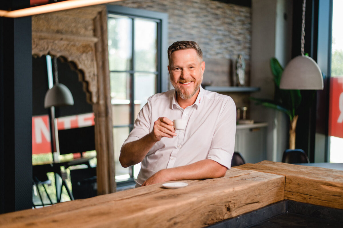 Christian Lorenz Fotograf Ein lächelnder Mann mit kurzen Haaren und Bart, der ein weißes Hemd trägt, hält eine kleine weiße Espressotasse, während er an einem hölzernen Tresen in einem modernen, stilvollen Raum mit Tageslicht steht. Christian Lorenz Fotograf Ein lächelnder Mann mit kurzen Haaren und Bart, der ein weißes Hemd trägt, hält eine kleine weiße Espressotasse, während er an einem hölzernen Tresen in einem modernen, stilvollen Raum mit Tageslicht steht.