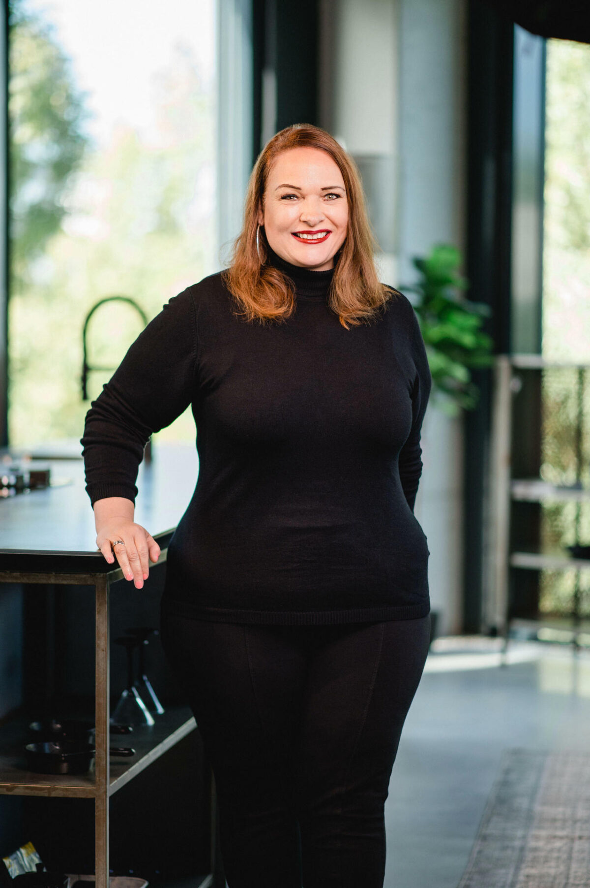 Christian Lorenz Fotograf Eine Frau mit hellbraunem Haar, die einen schwarzen Rollkragenpullover und eine schwarze Hose trägt, steht im Haus an einer Arbeitsplatte. Sie lächelt und stützt sich mit einer Hand auf die Arbeitsplatte. Im Hintergrund sind große Fenster und eine Pflanze zu sehen. Christian Lorenz Fotograf Eine Frau mit hellbraunem Haar, die einen schwarzen Rollkragenpullover und eine schwarze Hose trägt, steht im Haus an einer Arbeitsplatte. Sie lächelt und stützt sich mit einer Hand auf die Arbeitsplatte. Im Hintergrund sind große Fenster und eine Pflanze zu sehen.