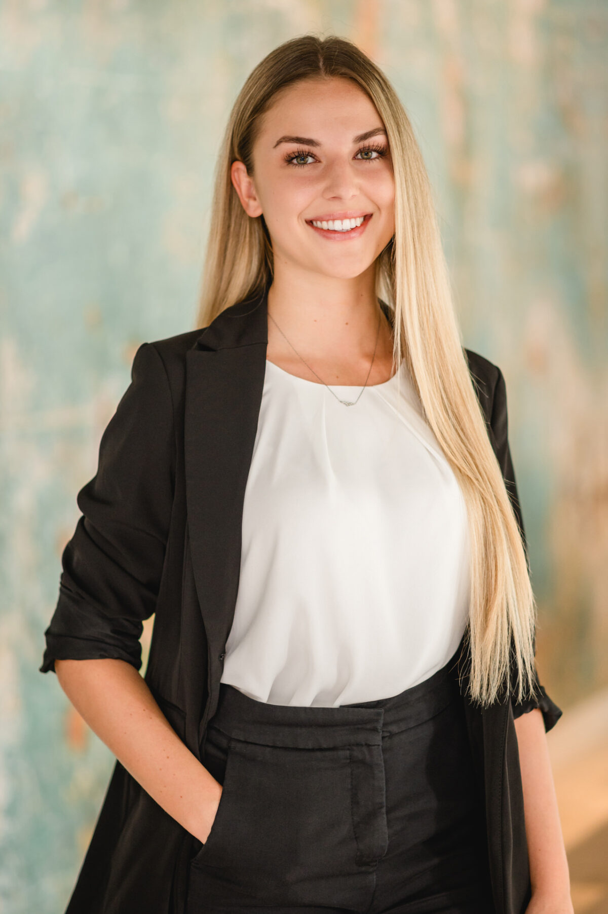 Christian Lorenz Fotograf Eine junge Frau mit langem blondem Haar steht lächelnd da und trägt einen schwarzen Blazer und eine Hose mit weißer Bluse. Sie hat eine Hand in der Tasche und posiert selbstbewusst vor einem sanft verschwommenen, grünlich strukturierten Hintergrund. Christian Lorenz Fotograf Eine junge Frau mit langem blondem Haar steht lächelnd da und trägt einen schwarzen Blazer und eine Hose mit weißer Bluse. Sie hat eine Hand in der Tasche und posiert selbstbewusst vor einem sanft verschwommenen, grünlich strukturierten Hintergrund.