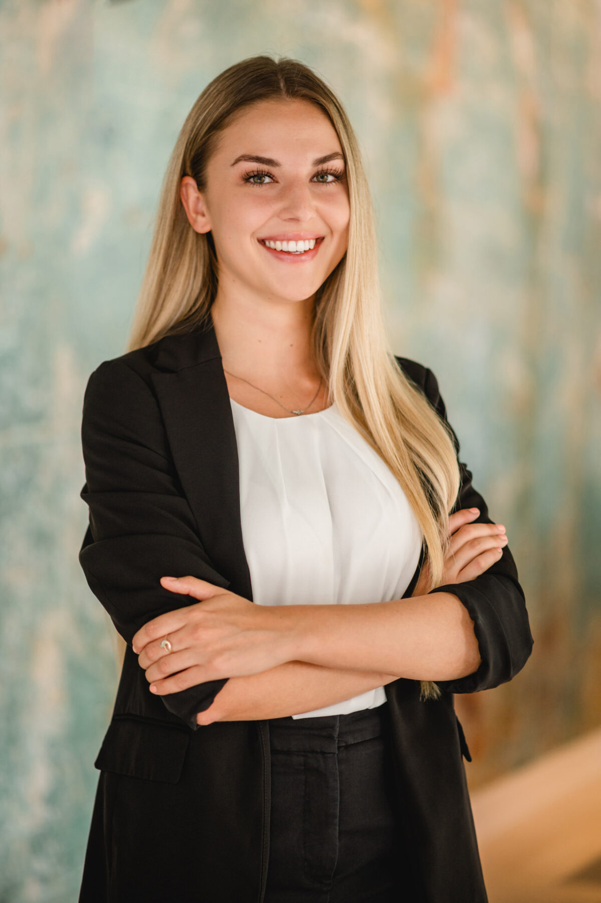 Christian Lorenz Fotograf Eine junge Frau mit langen blonden Haaren, schwarzem Blazer und weißem Oberteil steht lächelnd mit verschränkten Armen vor einem verschwommenen, hellblau-beigen Hintergrund. Christian Lorenz Fotograf Eine junge Frau mit langen blonden Haaren, schwarzem Blazer und weißem Oberteil steht lächelnd mit verschränkten Armen vor einem verschwommenen, hellblau-beigen Hintergrund.