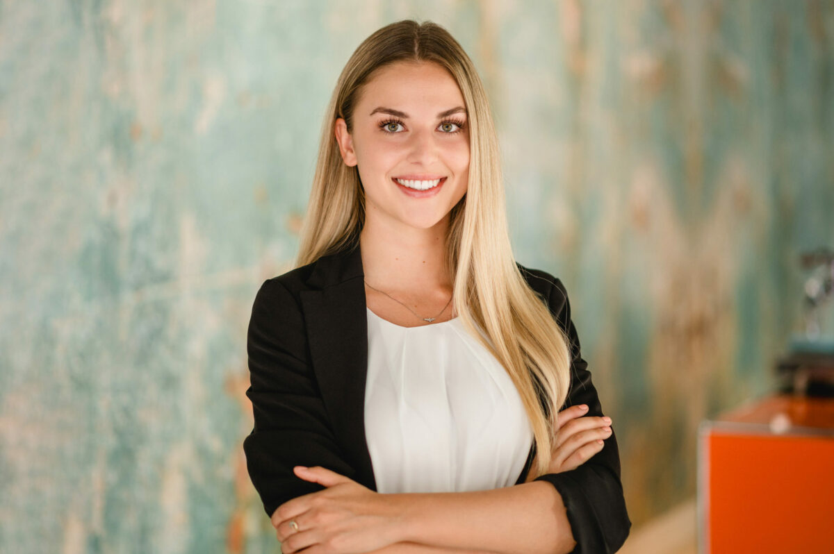 Christian Lorenz Fotograf Eine junge Frau mit langen blonden Haaren, die einen schwarzen Blazer und eine weiße Bluse trägt, steht lächelnd mit verschränkten Armen vor einem unscharfen blau-orangenen Hintergrund. Christian Lorenz Fotograf Eine junge Frau mit langen blonden Haaren, die einen schwarzen Blazer und eine weiße Bluse trägt, steht lächelnd mit verschränkten Armen vor einem unscharfen blau-orangenen Hintergrund.
