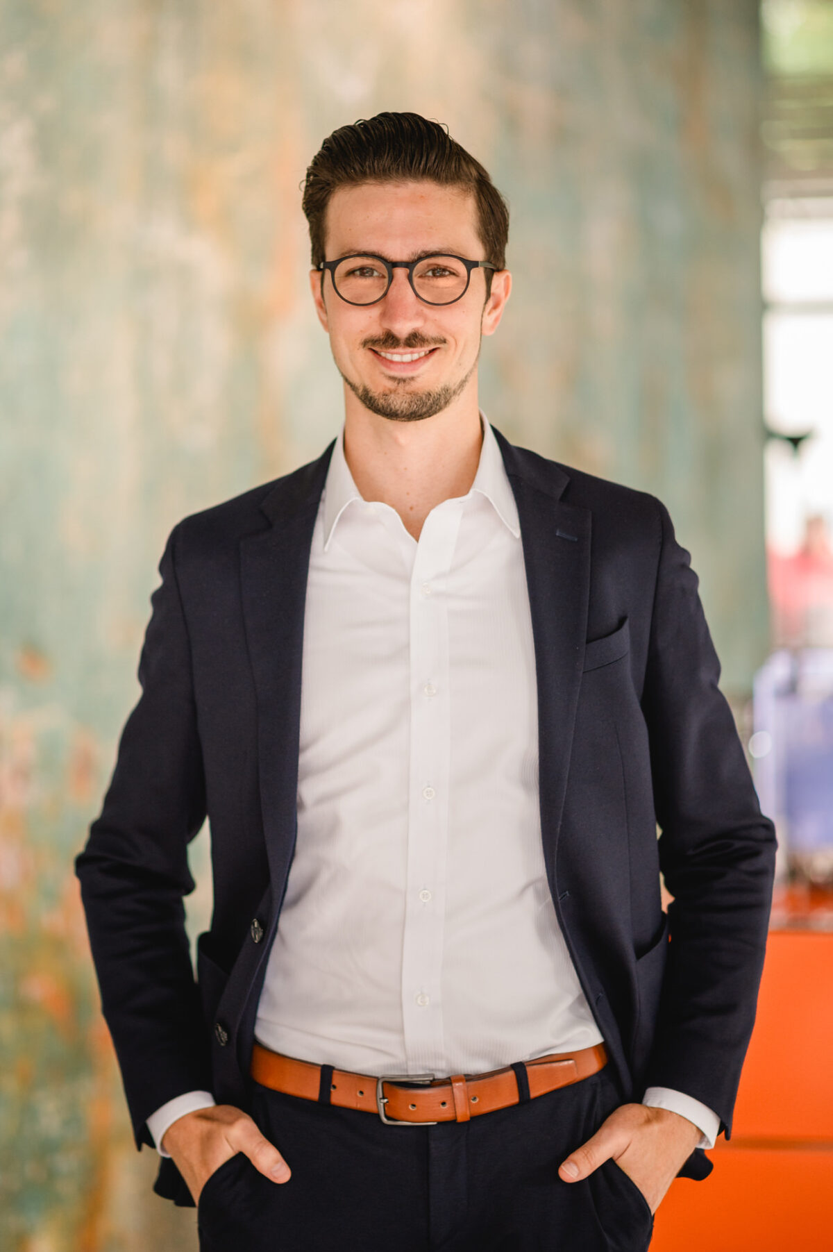 Christian Lorenz Fotograf Ein Mann mit Brille, marineblauem Anzug, weißem Hemd und braunem Gürtel steht mit den Händen in den Taschen und einem selbstbewussten Lächeln vor einem unscharfen, strukturierten Hintergrund. Christian Lorenz Fotograf Ein Mann mit Brille, marineblauem Anzug, weißem Hemd und braunem Gürtel steht mit den Händen in den Taschen und einem selbstbewussten Lächeln vor einem unscharfen, strukturierten Hintergrund.