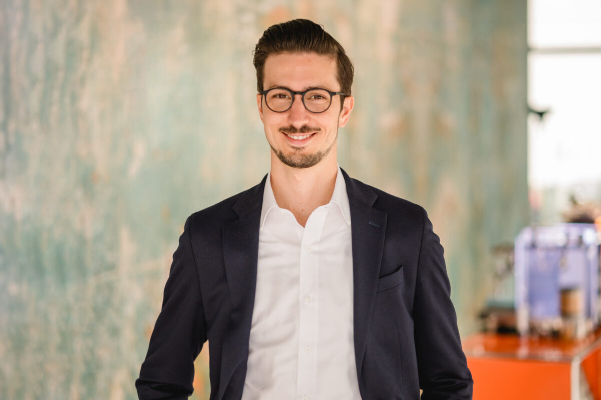 Christian Lorenz Fotograf Ein Mann mit kurzen braunen Haaren und Brille, der einen marineblauen Blazer und ein weißes Hemd trägt, steht lächelnd vor einer verschwommenen, strukturierten Wand mit einem orangefarbenen Objekt im Hintergrund. Christian Lorenz Fotograf Ein Mann mit kurzen braunen Haaren und Brille, der einen marineblauen Blazer und ein weißes Hemd trägt, steht lächelnd vor einer verschwommenen, strukturierten Wand mit einem orangefarbenen Objekt im Hintergrund.