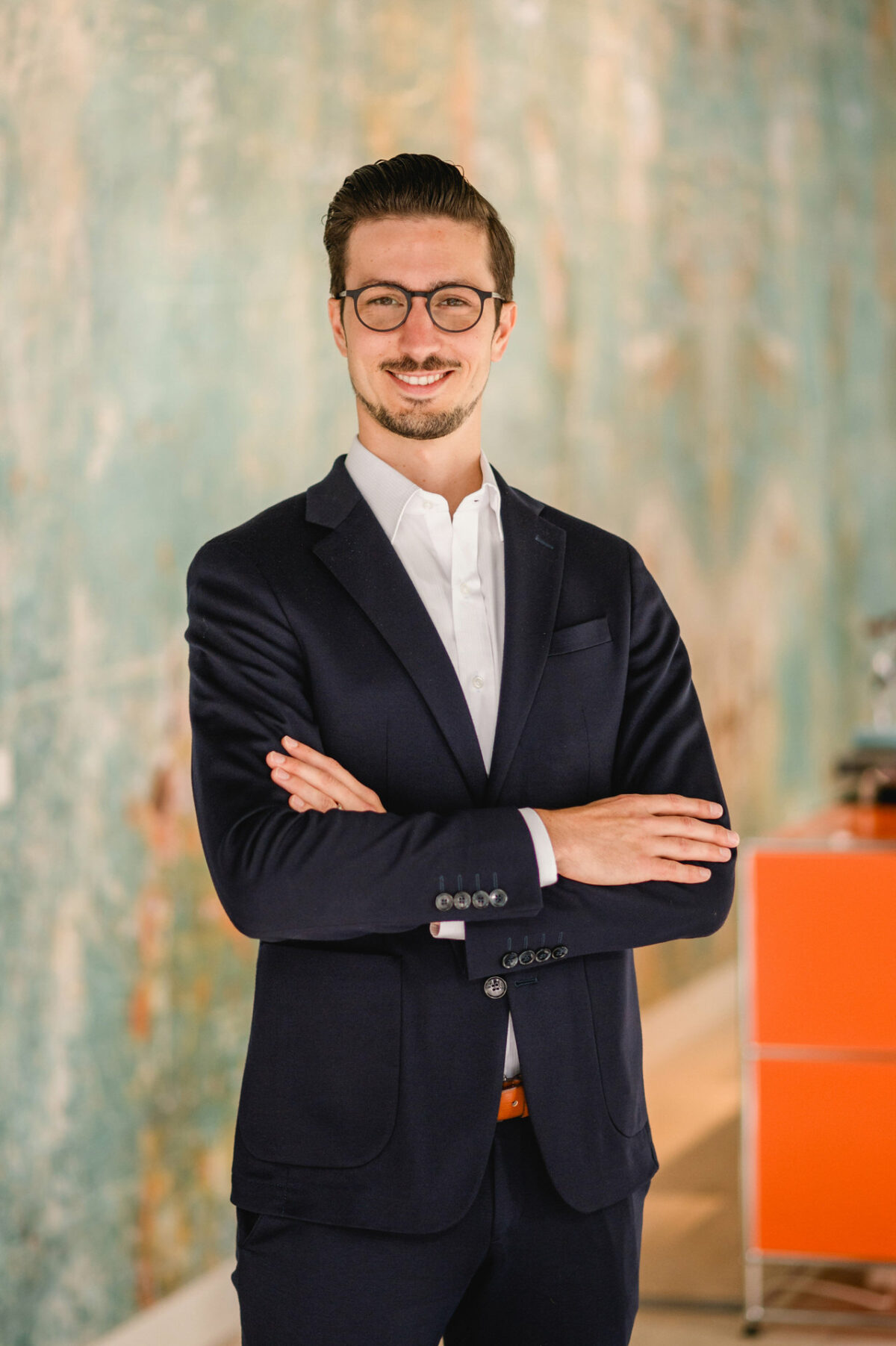 Christian Lorenz Fotograf Ein Mann in einem dunklen Anzug und mit Brille steht mit verschränkten Armen da und lächelt selbstbewusst. Der Hintergrund ist mit einer tealfarbenen, strukturierten Wand und einem orangefarbenen Möbelstück sanft verschwommen. Christian Lorenz Fotograf Ein Mann in einem dunklen Anzug und mit Brille steht mit verschränkten Armen da und lächelt selbstbewusst. Der Hintergrund ist mit einer tealfarbenen, strukturierten Wand und einem orangefarbenen Möbelstück sanft verschwommen.