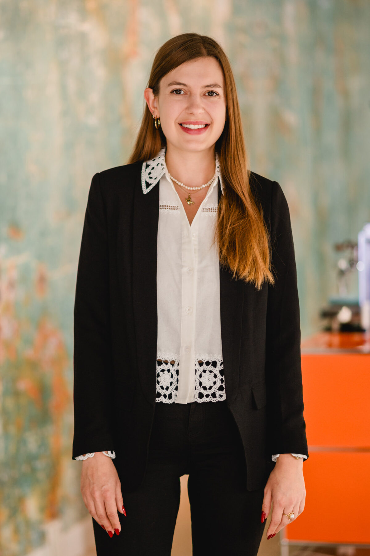 Christian Lorenz Fotograf Eine Frau mit langen braunen Haaren lächelt in die Kamera. Sie trägt einen schwarzen Blazer über einer weißen Bluse mit Spitzendetails und eine schwarze Hose. Der Hintergrund ist mit blauen und orangefarbenen Tönen sanft verschwommen. Christian Lorenz Fotograf Eine Frau mit langen braunen Haaren lächelt in die Kamera. Sie trägt einen schwarzen Blazer über einer weißen Bluse mit Spitzendetails und eine schwarze Hose. Der Hintergrund ist mit blauen und orangefarbenen Tönen sanft verschwommen.