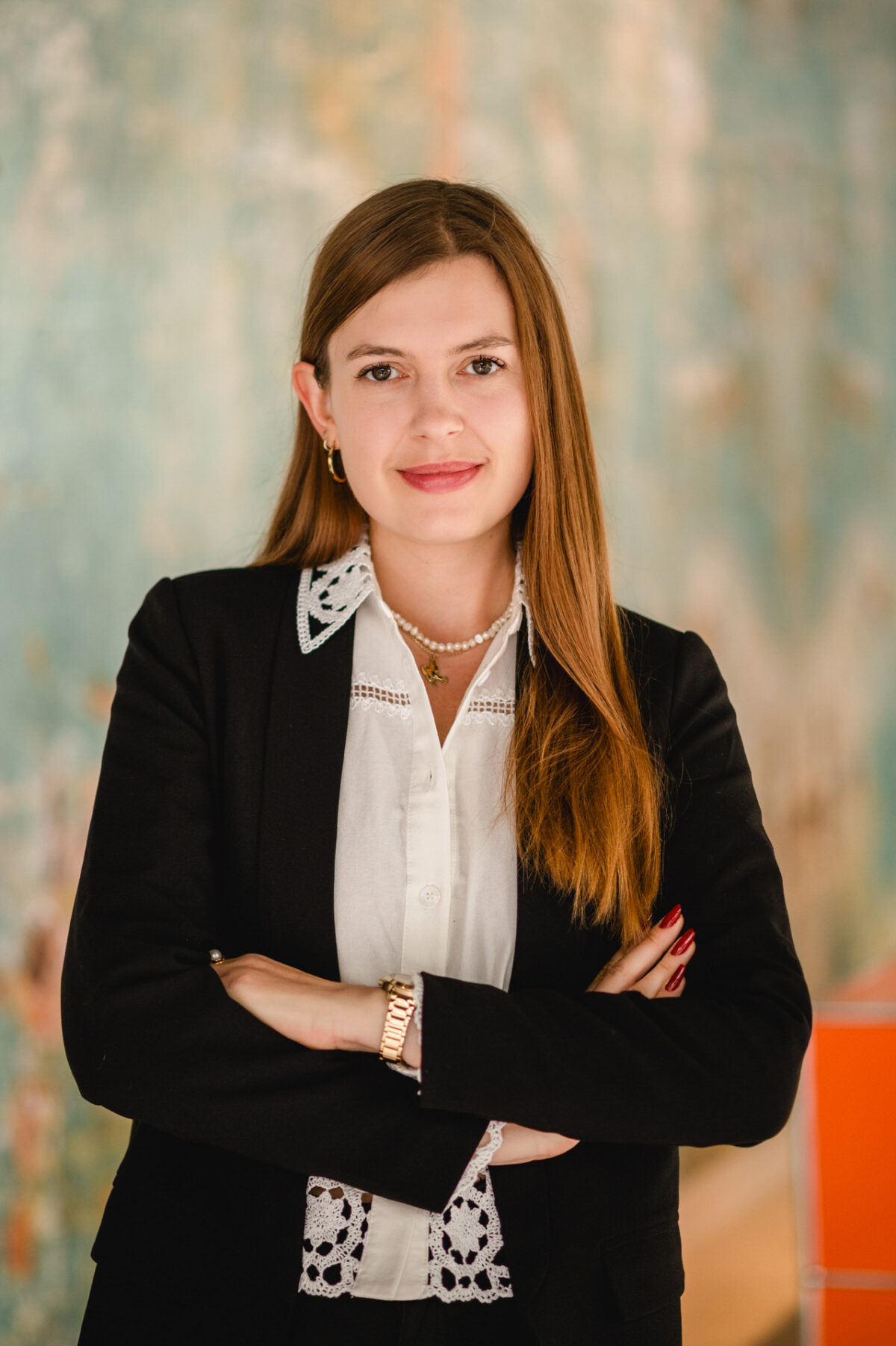 Christian Lorenz Fotograf Eine junge Frau mit langen braunen Haaren, die einen schwarzen Blazer über einer weißen Bluse trägt, steht mit verschränkten Armen und lächelt selbstbewusst in die Kamera. Der Hintergrund ist mit blauen und orangefarbenen Tönen sanft verschwommen. Christian Lorenz Fotograf Eine junge Frau mit langen braunen Haaren, die einen schwarzen Blazer über einer weißen Bluse trägt, steht mit verschränkten Armen und lächelt selbstbewusst in die Kamera. Der Hintergrund ist mit blauen und orangefarbenen Tönen sanft verschwommen.