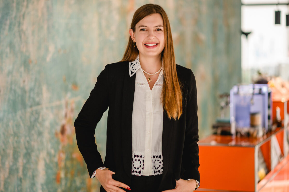 Christian Lorenz Fotograf Eine lächelnde Frau mit langen braunen Haaren steht in einem Haus, trägt einen schwarzen Blazer über einer weißen Bluse und hat die Hände in die Hüften gestemmt. Im Hintergrund sind eine strukturierte Wand und orangefarbene Möbel zu sehen. Christian Lorenz Fotograf Eine lächelnde Frau mit langen braunen Haaren steht in einem Haus, trägt einen schwarzen Blazer über einer weißen Bluse und hat die Hände in die Hüften gestemmt. Im Hintergrund sind eine strukturierte Wand und orangefarbene Möbel zu sehen.