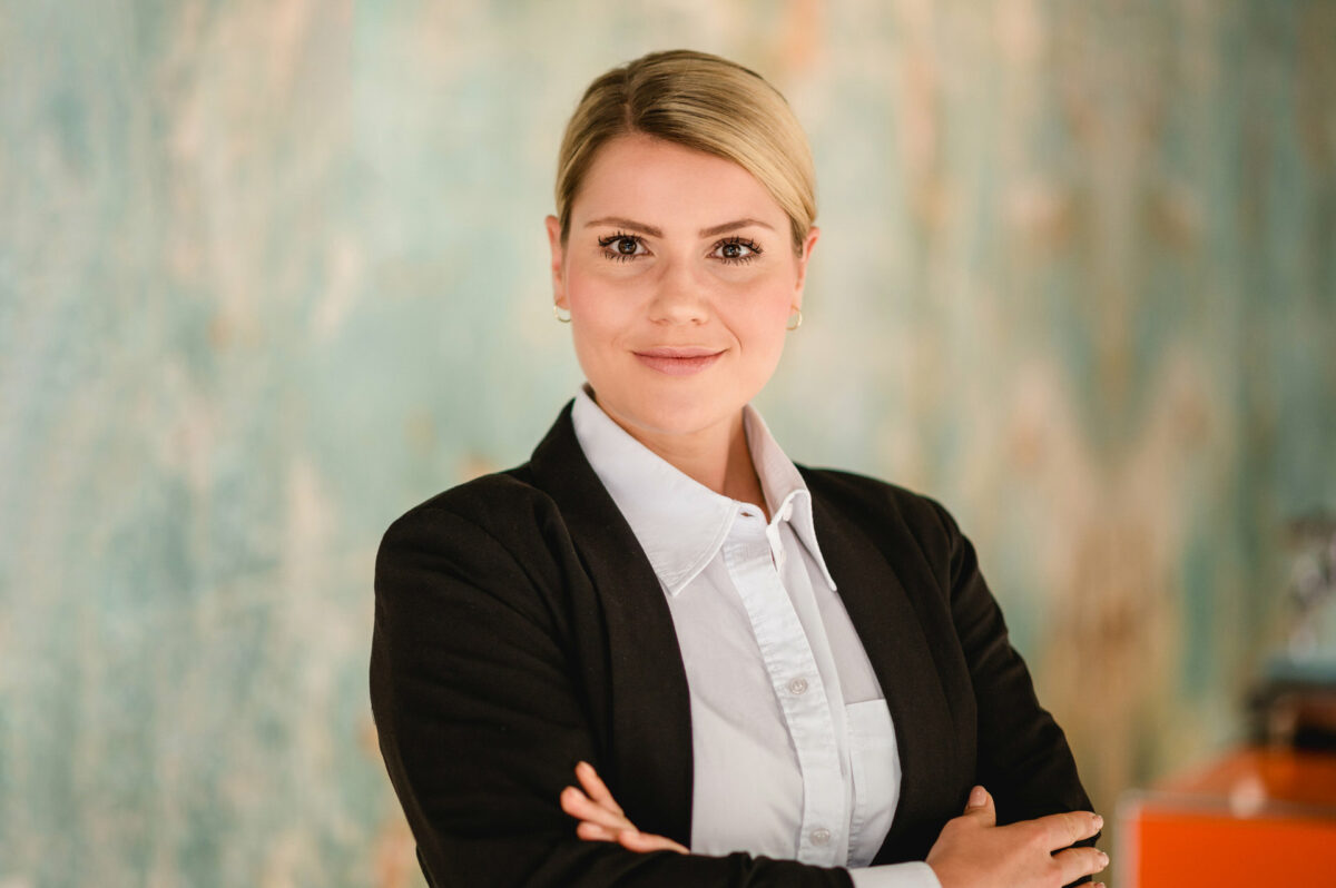 Christian Lorenz Fotograf Eine selbstbewusste Frau mit blondem Haar in einem gepflegten Dutt, die einen schwarzen Blazer über einem weißen Button-up-Hemd trägt, steht mit verschränkten Armen da und lächelt in die Kamera vor einem unscharfen, blau getönten Hintergrund. Christian Lorenz Fotograf Eine selbstbewusste Frau mit blondem Haar in einem gepflegten Dutt, die einen schwarzen Blazer über einem weißen Button-up-Hemd trägt, steht mit verschränkten Armen da und lächelt in die Kamera vor einem unscharfen, blau getönten Hintergrund.