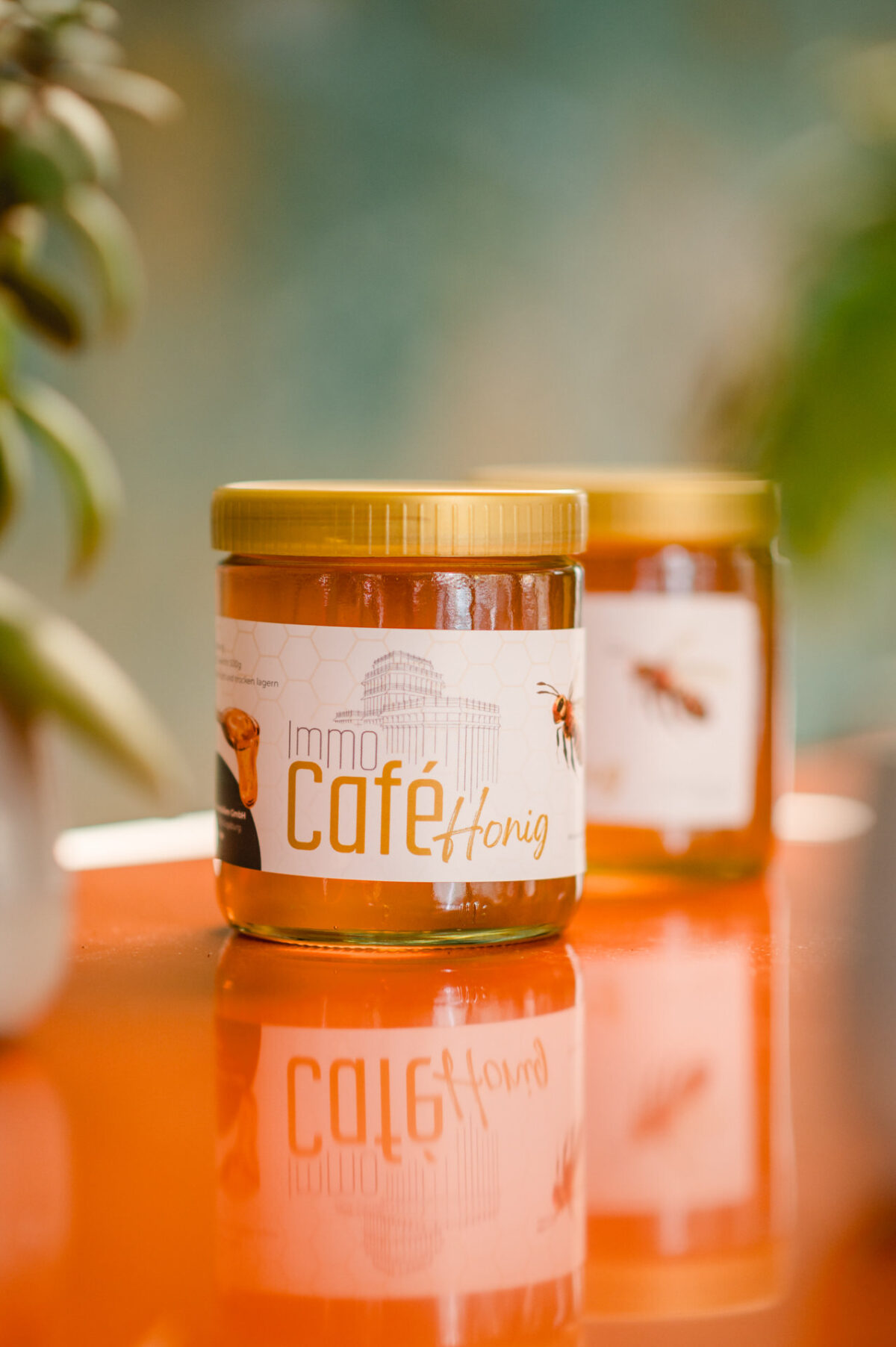 Christian Lorenz Fotograf Ein Glas Honig mit der Aufschrift "Immo Café Honig" und einem gelben Deckel steht auf einer orangefarbenen Fläche und spiegelt sich darunter. Ein weiteres Glas steht unscharf im Hintergrund. An den Rändern ist Grünzeug zu sehen. Christian Lorenz Fotograf Ein Glas Honig mit der Aufschrift "Immo Café Honig" und einem gelben Deckel steht auf einer orangefarbenen Fläche und spiegelt sich darunter. Ein weiteres Glas steht unscharf im Hintergrund. An den Rändern ist Grünzeug zu sehen.
