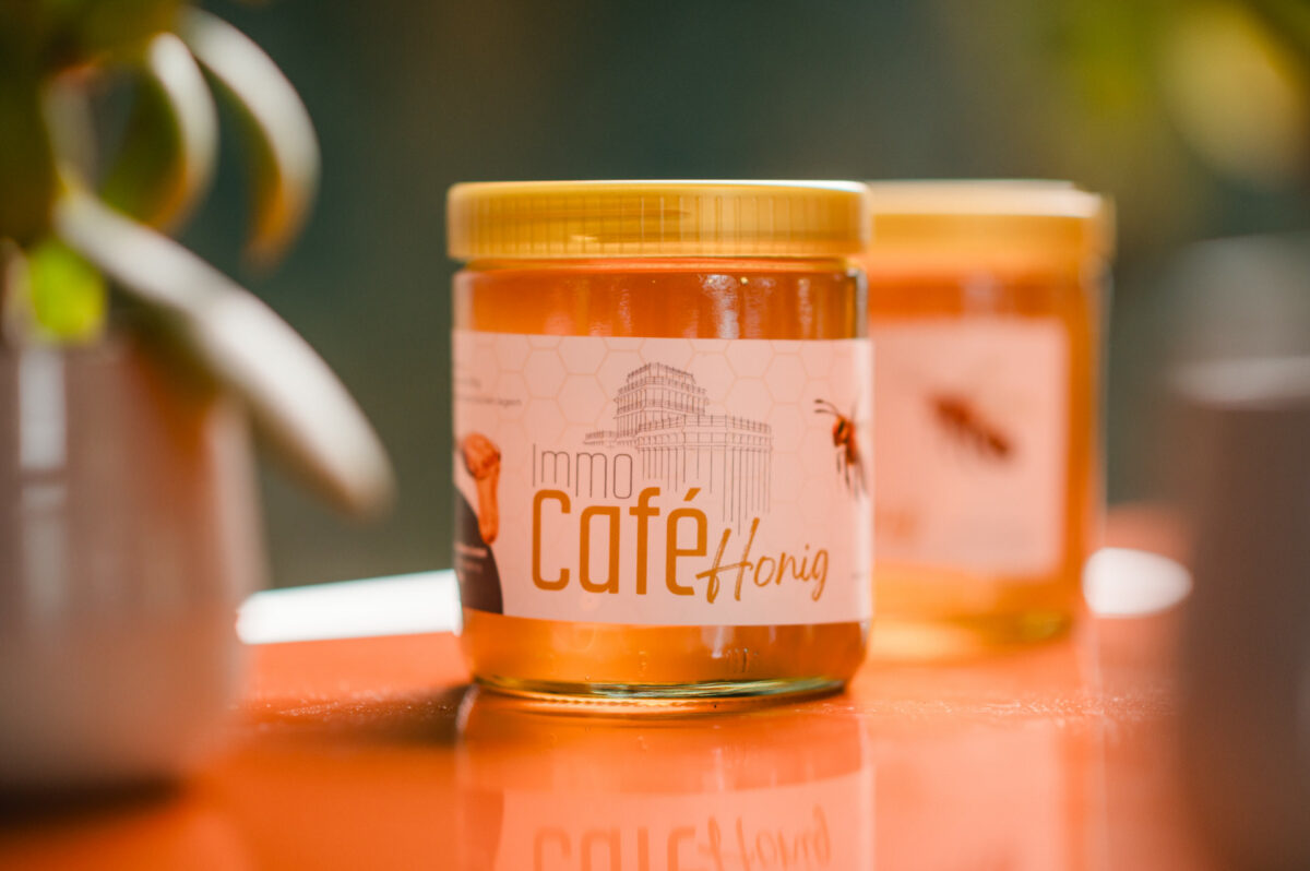 Christian Lorenz Fotograf Ein Glas Honig mit der Aufschrift "immo Café Honig" steht auf einer orangefarbenen Fläche, während ein anderes Glas und eine grüne Pflanze im Hintergrund verschwimmen. Das Etikett zeigt eine Biene, ein Wabenmuster und den Umriss eines Gebäudes. Christian Lorenz Fotograf Ein Glas Honig mit der Aufschrift "immo Café Honig" steht auf einer orangefarbenen Fläche, während ein anderes Glas und eine grüne Pflanze im Hintergrund verschwimmen. Das Etikett zeigt eine Biene, ein Wabenmuster und den Umriss eines Gebäudes.