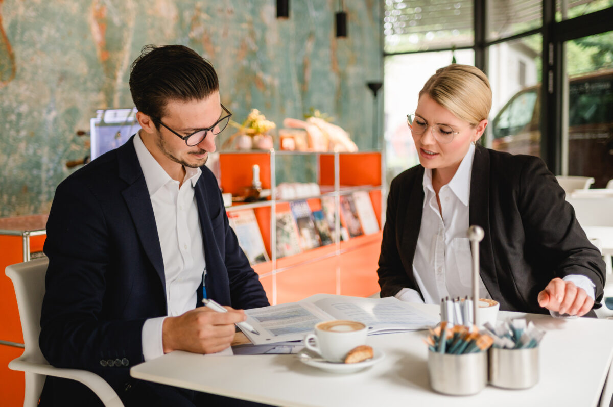 Christian Lorenz Fotograf Zwei Personen in Geschäftskleidung sitzen an einem Tisch in einem modernen Büro und prüfen gemeinsam Dokumente mit Kaffeetassen vor ihnen. Im Hintergrund sind Regale mit Zeitschriften und eine dekorative Wand zu sehen. Christian Lorenz Fotograf Zwei Personen in Geschäftskleidung sitzen an einem Tisch in einem modernen Büro und prüfen gemeinsam Dokumente mit Kaffeetassen vor ihnen. Im Hintergrund sind Regale mit Zeitschriften und eine dekorative Wand zu sehen.