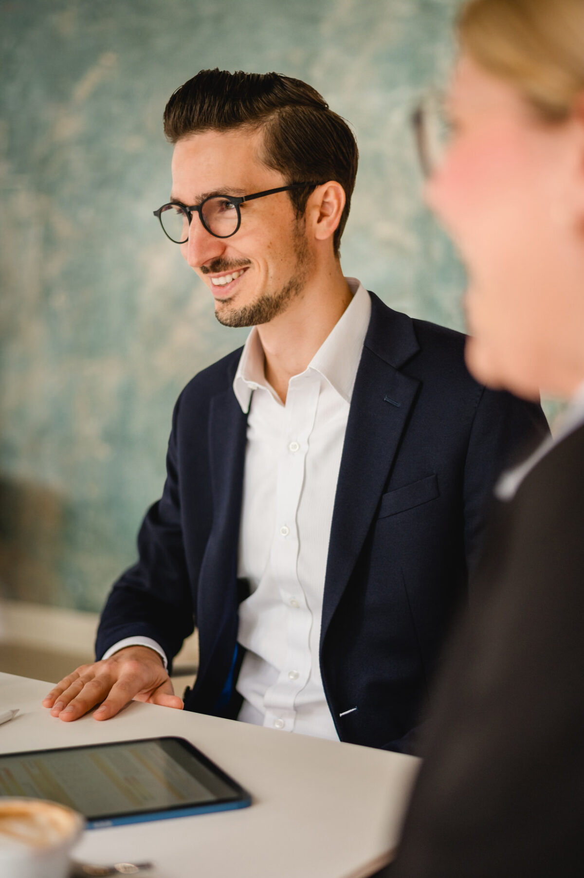 Christian Lorenz Fotograf Ein Mann in einem marineblauen Anzug und weißem Hemd sitzt an einem Tisch, lächelt und blickt zur Seite. Er trägt eine Brille, und vor ihm steht ein Tablet mit Dokumenten auf dem Bildschirm. Eine weitere Person ist unscharf im Vordergrund zu erkennen. Christian Lorenz Fotograf Ein Mann in einem marineblauen Anzug und weißem Hemd sitzt an einem Tisch, lächelt und blickt zur Seite. Er trägt eine Brille, und vor ihm steht ein Tablet mit Dokumenten auf dem Bildschirm. Eine weitere Person ist unscharf im Vordergrund zu erkennen.