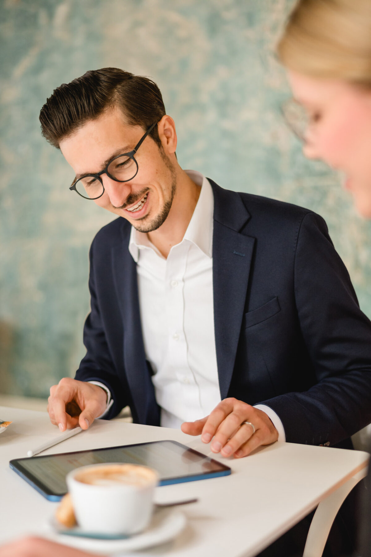 Christian Lorenz Fotograf Ein Mann in einem marineblauen Anzug und mit Brille lächelt, während er an einem Tisch in einem Café ein Tablet benutzt, mit einer Tasse Kaffee und einer anderen Person im Vordergrund. Christian Lorenz Fotograf Ein Mann in einem marineblauen Anzug und mit Brille lächelt, während er an einem Tisch in einem Café ein Tablet benutzt, mit einer Tasse Kaffee und einer anderen Person im Vordergrund.