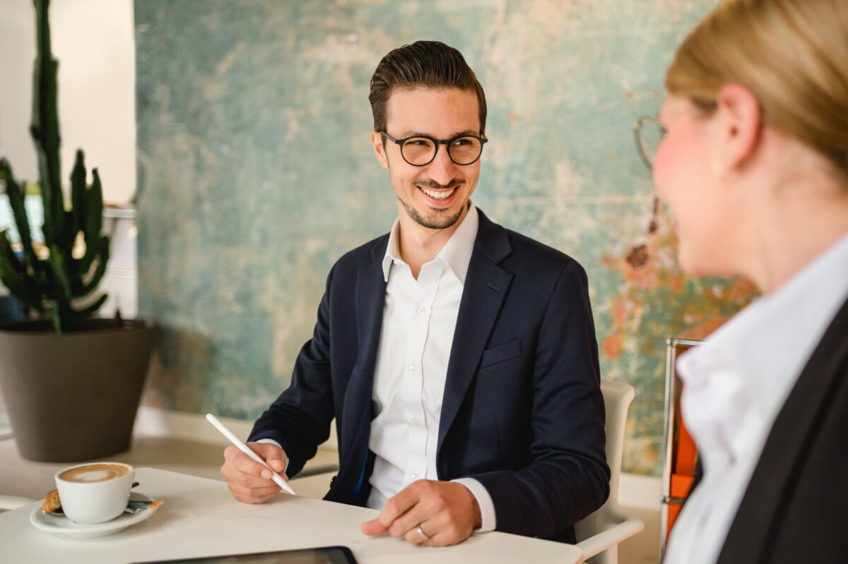 Christian Lorenz Fotograf Ein Mann im Anzug und mit Brille lächelt, während er einen Stift in der Hand hält und mit einer Frau in einer Büroumgebung spricht. Auf dem Tisch stehen eine Tasse Kaffee und ein digitales Tablet; im Hintergrund steht ein großer Kaktus in einem Topf. Christian Lorenz Fotograf Ein Mann im Anzug und mit Brille lächelt, während er einen Stift in der Hand hält und mit einer Frau in einer Büroumgebung spricht. Auf dem Tisch stehen eine Tasse Kaffee und ein digitales Tablet; im Hintergrund steht ein großer Kaktus in einem Topf.