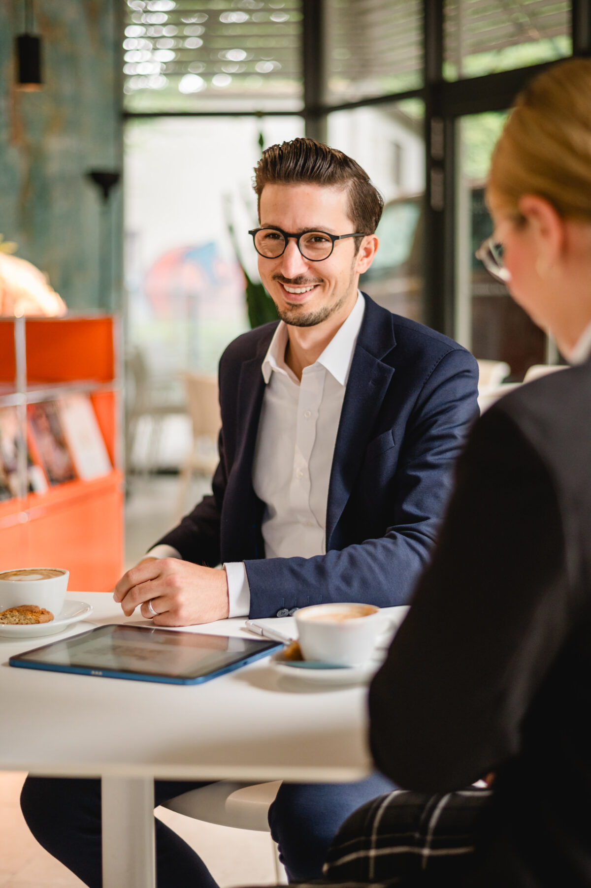 Christian Lorenz Fotograf Ein Mann im Anzug und mit Brille sitzt an einem Tisch mit Kaffee und Gebäck und lächelt eine andere Person in einem hellen, modernen Café an. Auf dem Tisch stehen ein Tablet und ein Teller. Christian Lorenz Fotograf Ein Mann im Anzug und mit Brille sitzt an einem Tisch mit Kaffee und Gebäck und lächelt eine andere Person in einem hellen, modernen Café an. Auf dem Tisch stehen ein Tablet und ein Teller.