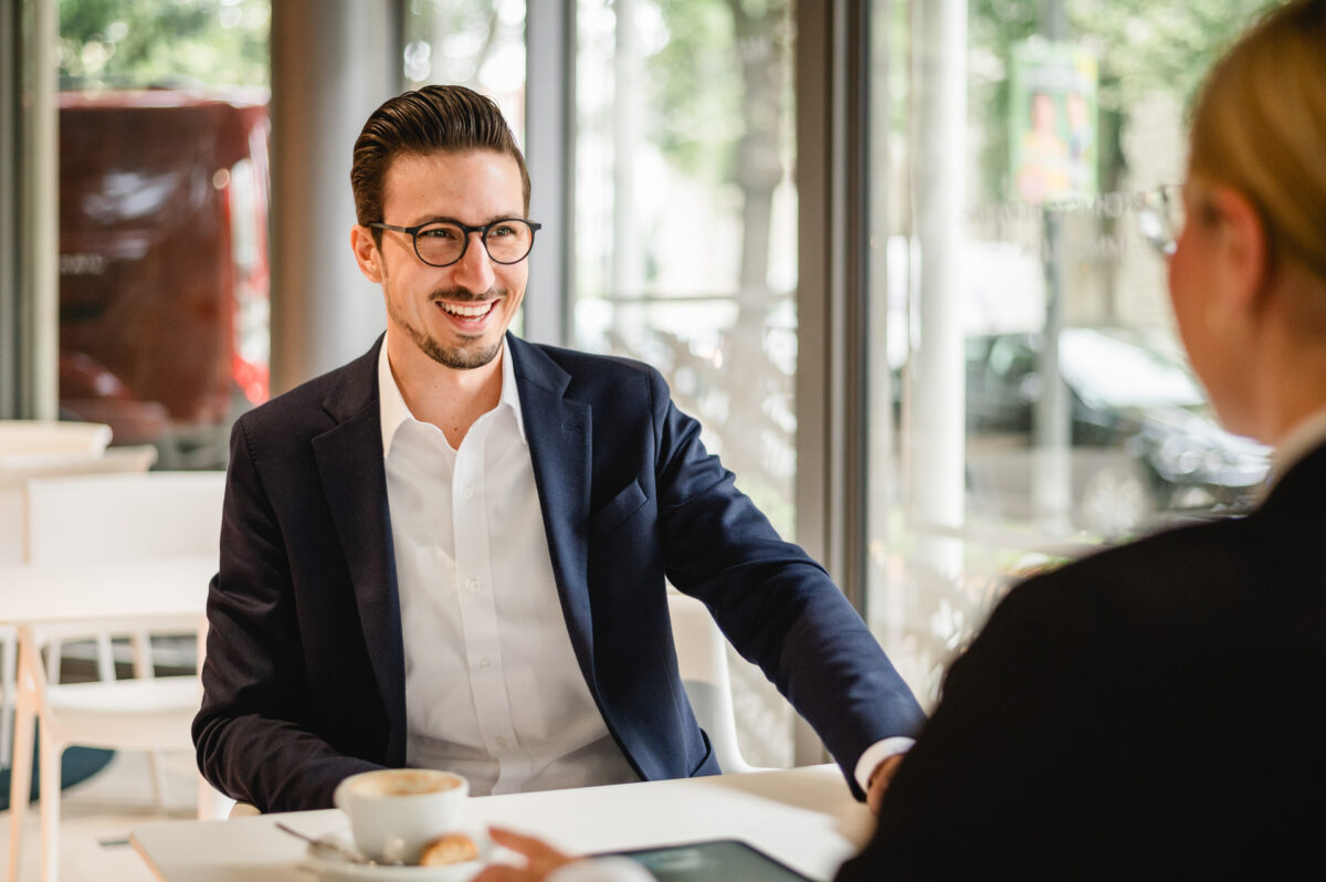 Christian Lorenz Fotograf Ein Mann im Anzug und mit Brille sitzt an einem Tisch in einem hellen Café, lächelt und spricht mit einer anderen Person. Auf dem Tisch vor ihm stehen eine Tasse Kaffee und ein Gebäckstück. Christian Lorenz Fotograf Ein Mann im Anzug und mit Brille sitzt an einem Tisch in einem hellen Café, lächelt und spricht mit einer anderen Person. Auf dem Tisch vor ihm stehen eine Tasse Kaffee und ein Gebäckstück.