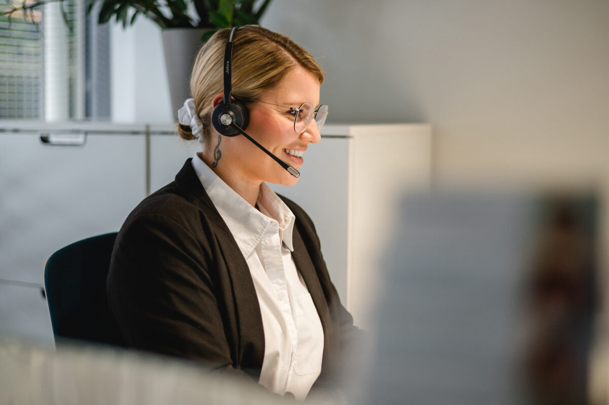 Christian Lorenz Fotograf Eine Frau mit Brille und Headset sitzt an einem Schreibtisch in einem Büro und lächelt, während sie arbeitet, mit einer Pflanze und Schränken im Hintergrund. Christian Lorenz Fotograf Eine Frau mit Brille und Headset sitzt an einem Schreibtisch in einem Büro und lächelt, während sie arbeitet, mit einer Pflanze und Schränken im Hintergrund.