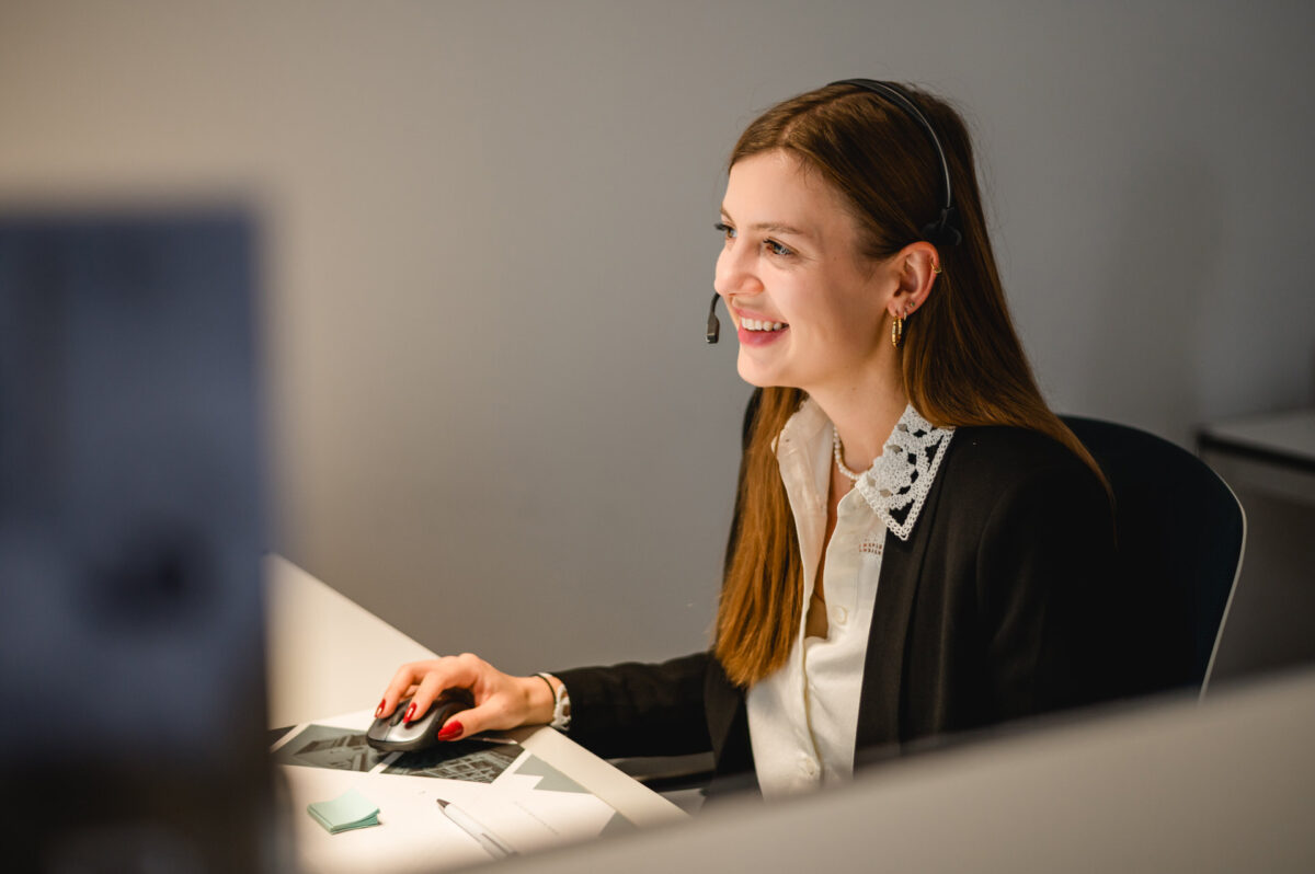 Christian Lorenz Fotograf Eine Frau, die ein Headset und Geschäftskleidung trägt, lächelt, während sie an einem Schreibtisch mit einer Computermaus und Dokumenten in einer Büroumgebung arbeitet. Christian Lorenz Fotograf Eine Frau, die ein Headset und Geschäftskleidung trägt, lächelt, während sie an einem Schreibtisch mit einer Computermaus und Dokumenten in einer Büroumgebung arbeitet.