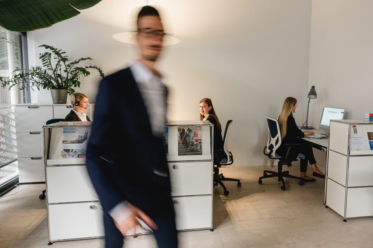 Christian Lorenz Fotograf Ein verschwommener Mann im Anzug geht an drei Frauen vorbei, die an Schreibtischen in einem modernen, hellen Büro mit Pflanzen und minimalistischer Einrichtung arbeiten. Christian Lorenz Fotograf Ein verschwommener Mann im Anzug geht an drei Frauen vorbei, die an Schreibtischen in einem modernen, hellen Büro mit Pflanzen und minimalistischer Einrichtung arbeiten.