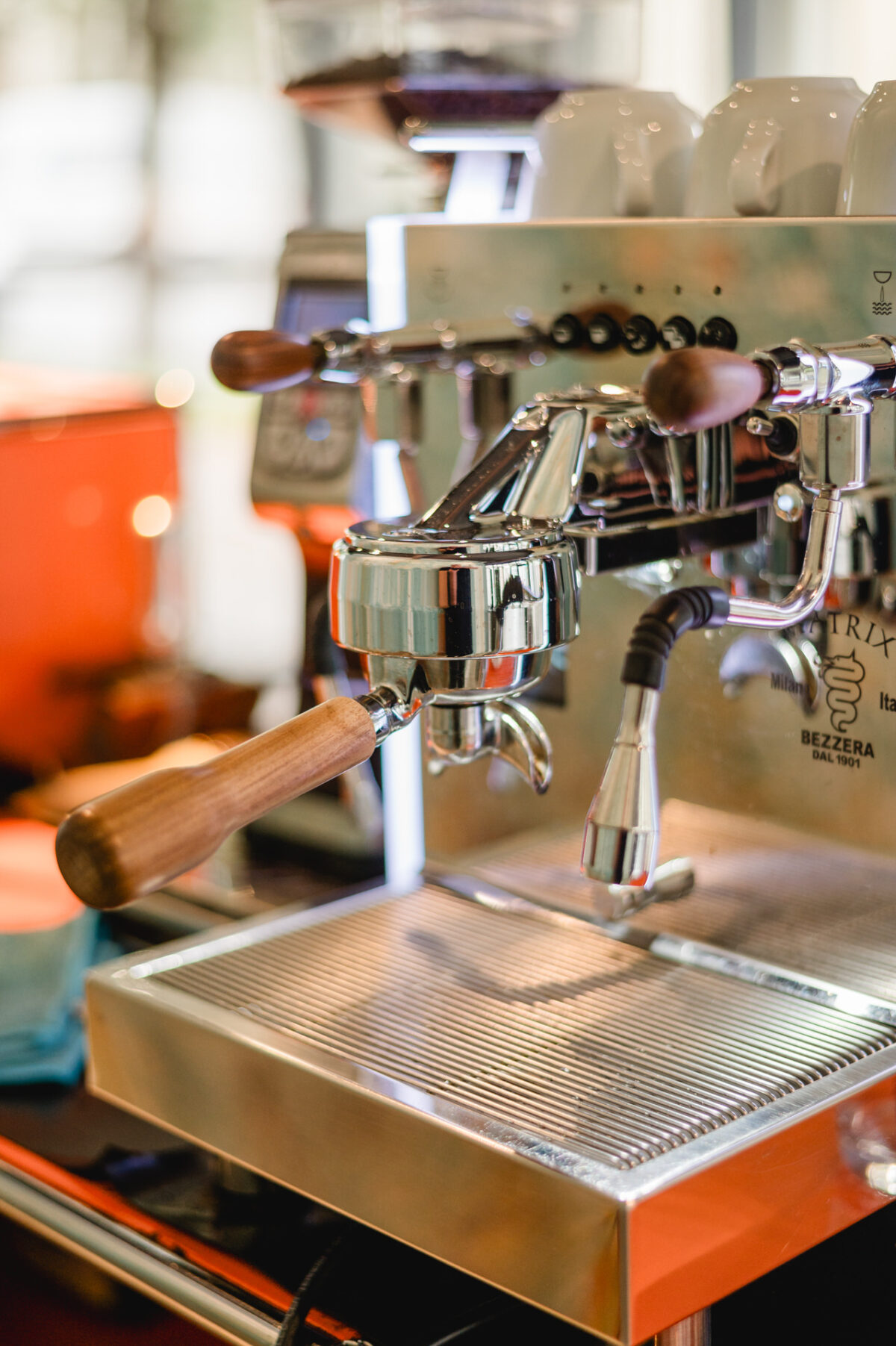 Christian Lorenz Fotograf Nahaufnahme einer glänzenden Espressomaschine mit Holzgriffen, die mit einem Siebträger und einer Dampfdüse ausgestattet ist und in einem hell erleuchteten Café auf einer Metallabtropfschale steht. Christian Lorenz Fotograf Nahaufnahme einer glänzenden Espressomaschine mit Holzgriffen, die mit einem Siebträger und einer Dampfdüse ausgestattet ist und in einem hell erleuchteten Café auf einer Metallabtropfschale steht.